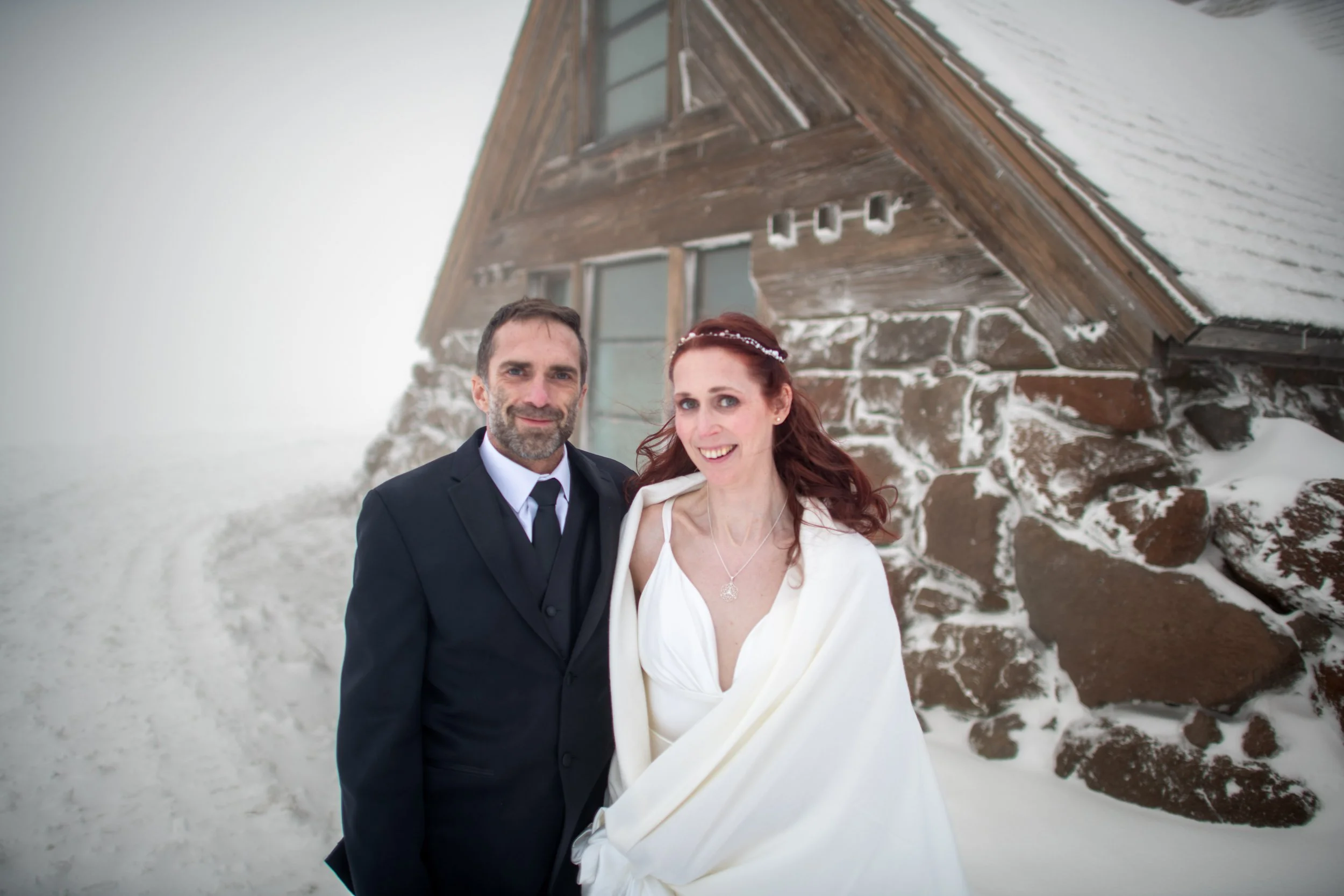 A man in a dark suit and a woman in a white wedding dress standing outside on a snow-covered area with a wooden house in the background.