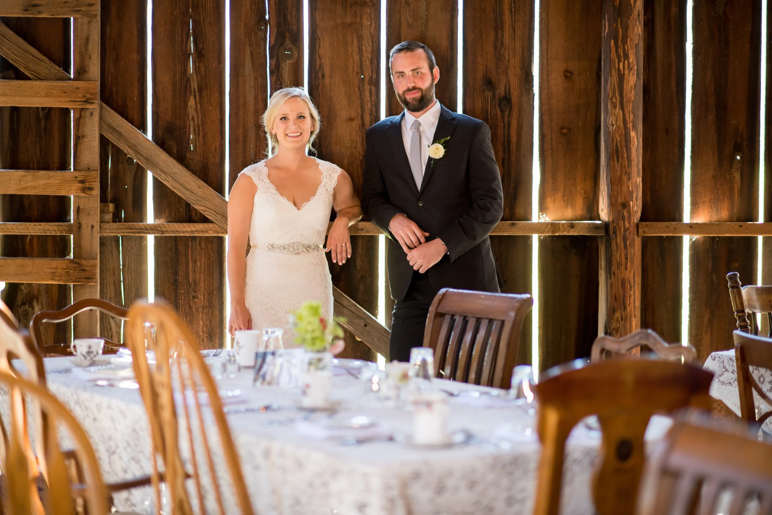 A bride and groom standing together inside a rustic wooden barn, celebrating their wedding. The bride wears a white lace gown and the groom wears a black suit with a white shirt and tie. They are smiling and holding hands, with tables set for guests 