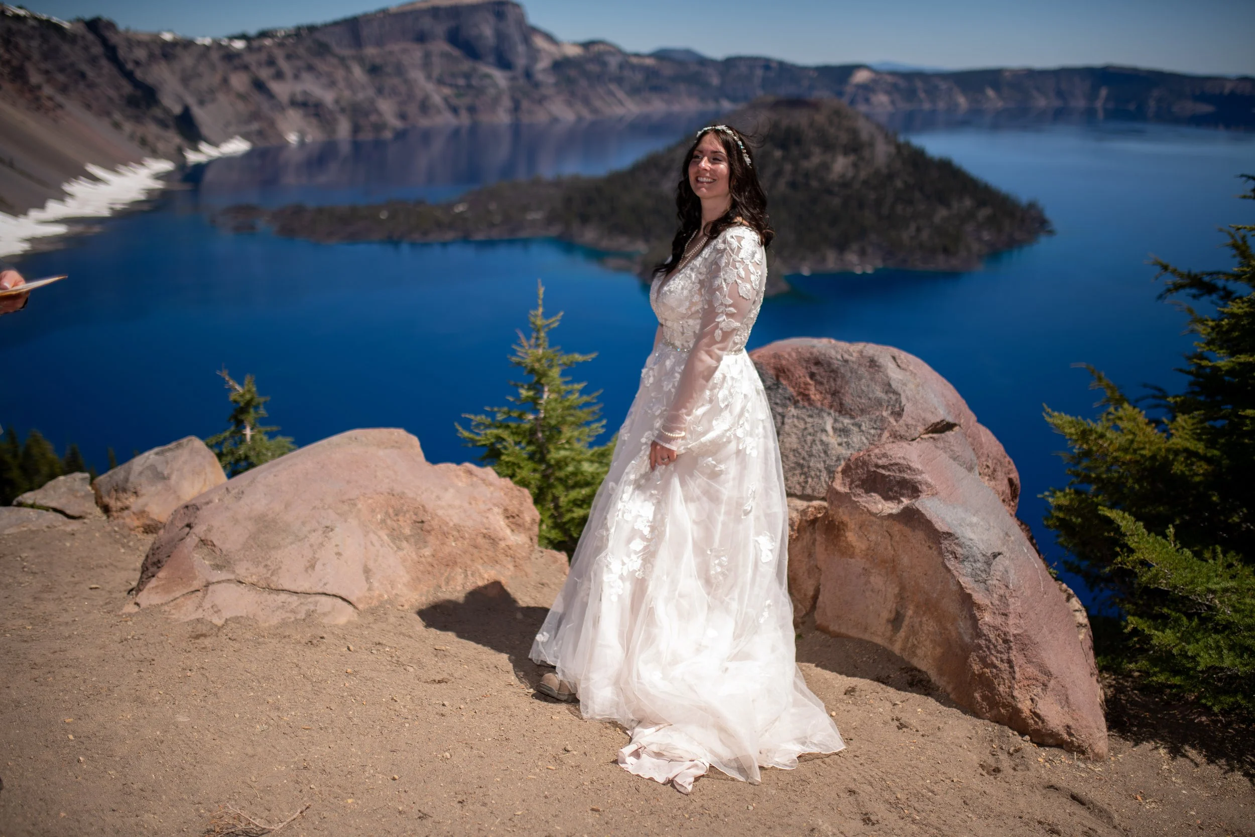 A woman dressed in a wedding gown stands on rocky terrain near a lake with mountains in the background. She is smiling and facing slightly to the side.