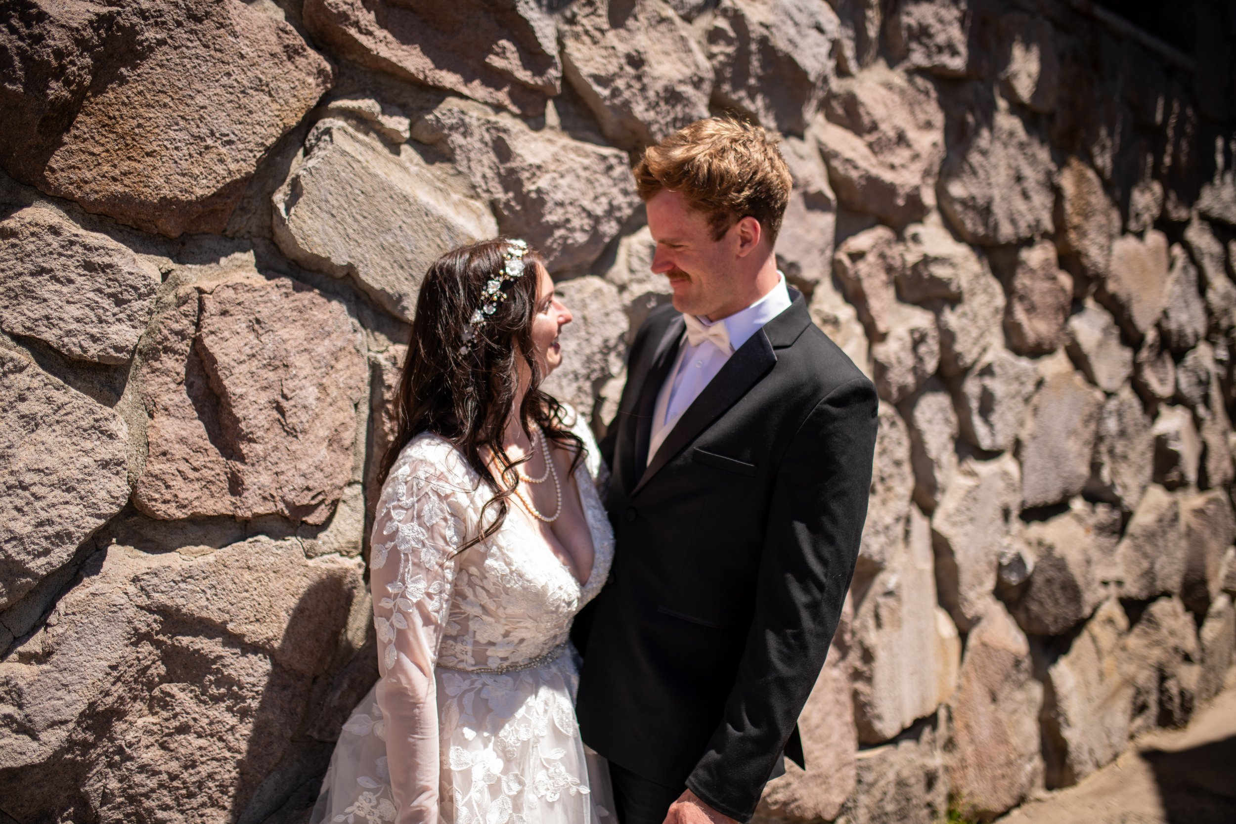 A bride and groom standing close together against a stone wall, smiling and gazing at each other outdoors on a sunny day.