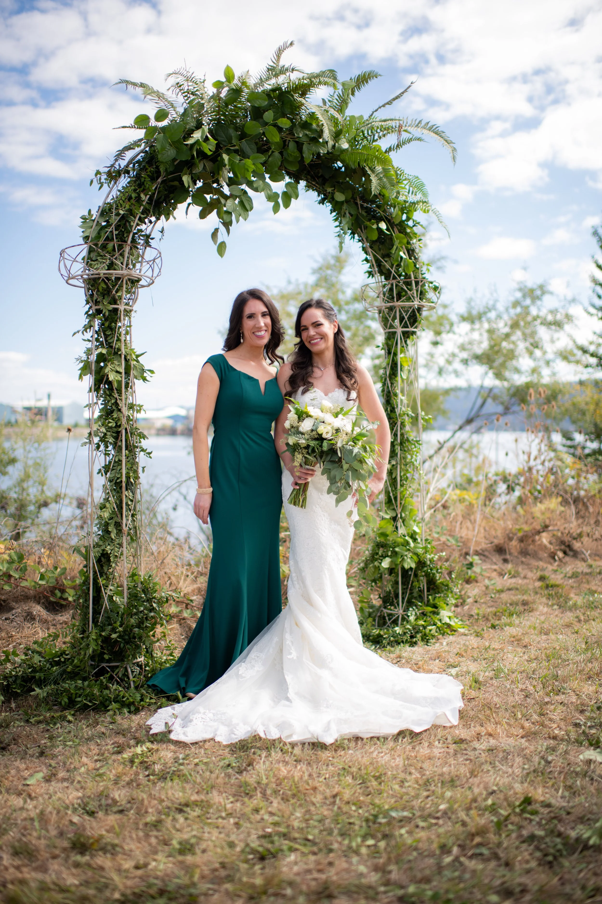 Two women standing under a decorative arch made of greenery, with a background of lake and sky. One woman wears a white wedding dress holding a bouquet, and the other wears a teal dress.