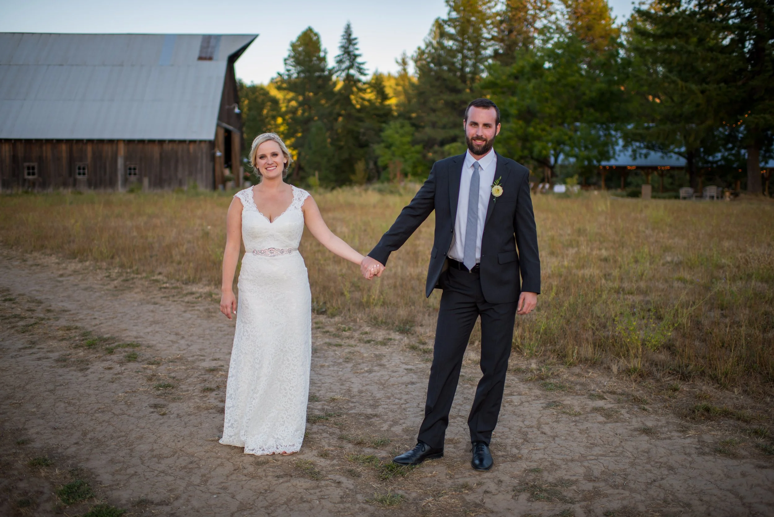 A bride in a white lace wedding dress holding hands with a groom in a black suit and gray tie outdoors during sunset with trees and a rustic barn in the background.