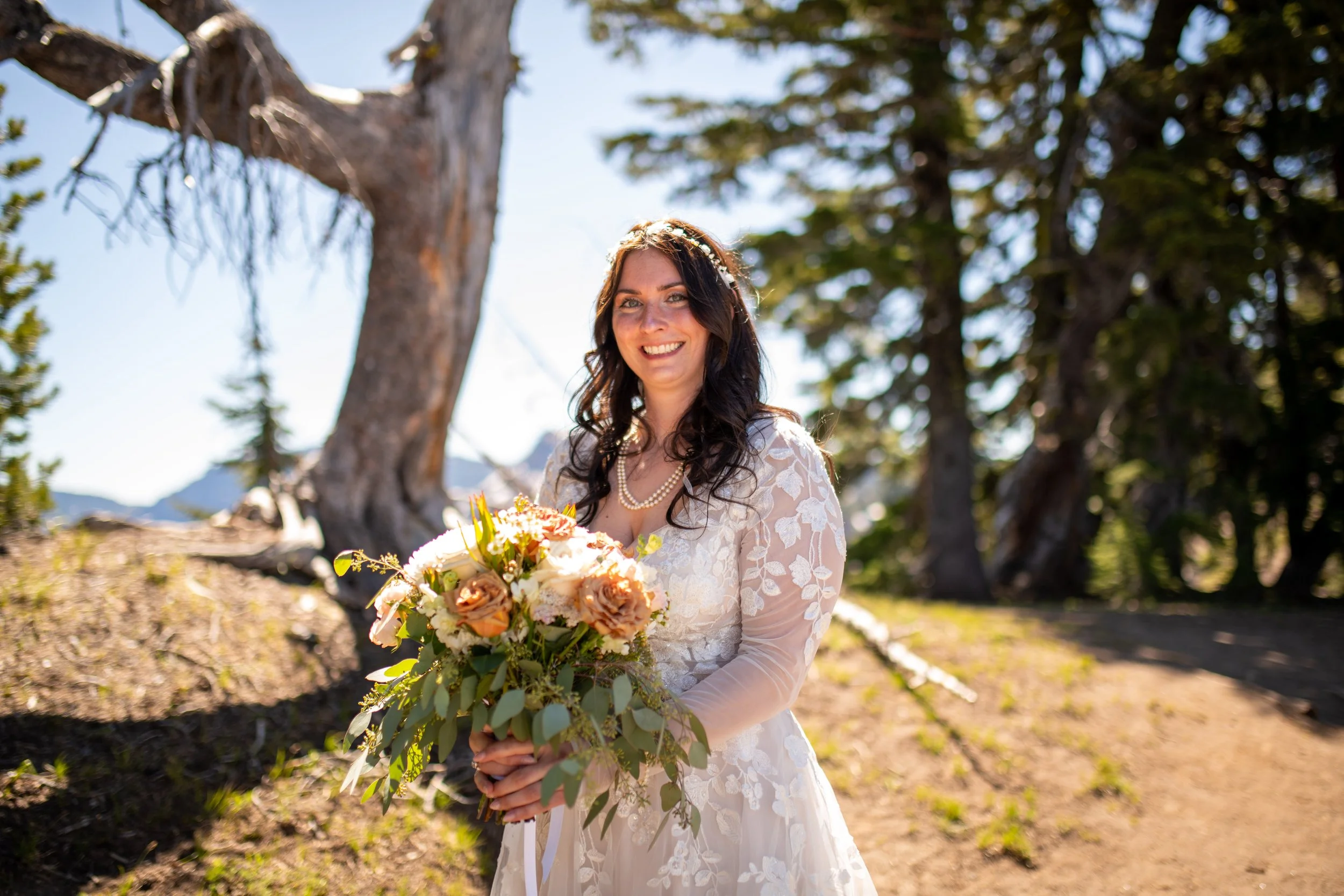 A woman in a lace wedding dress holding a bouquet of flowers outdoors on a sunny day, with trees and mountains in the background.