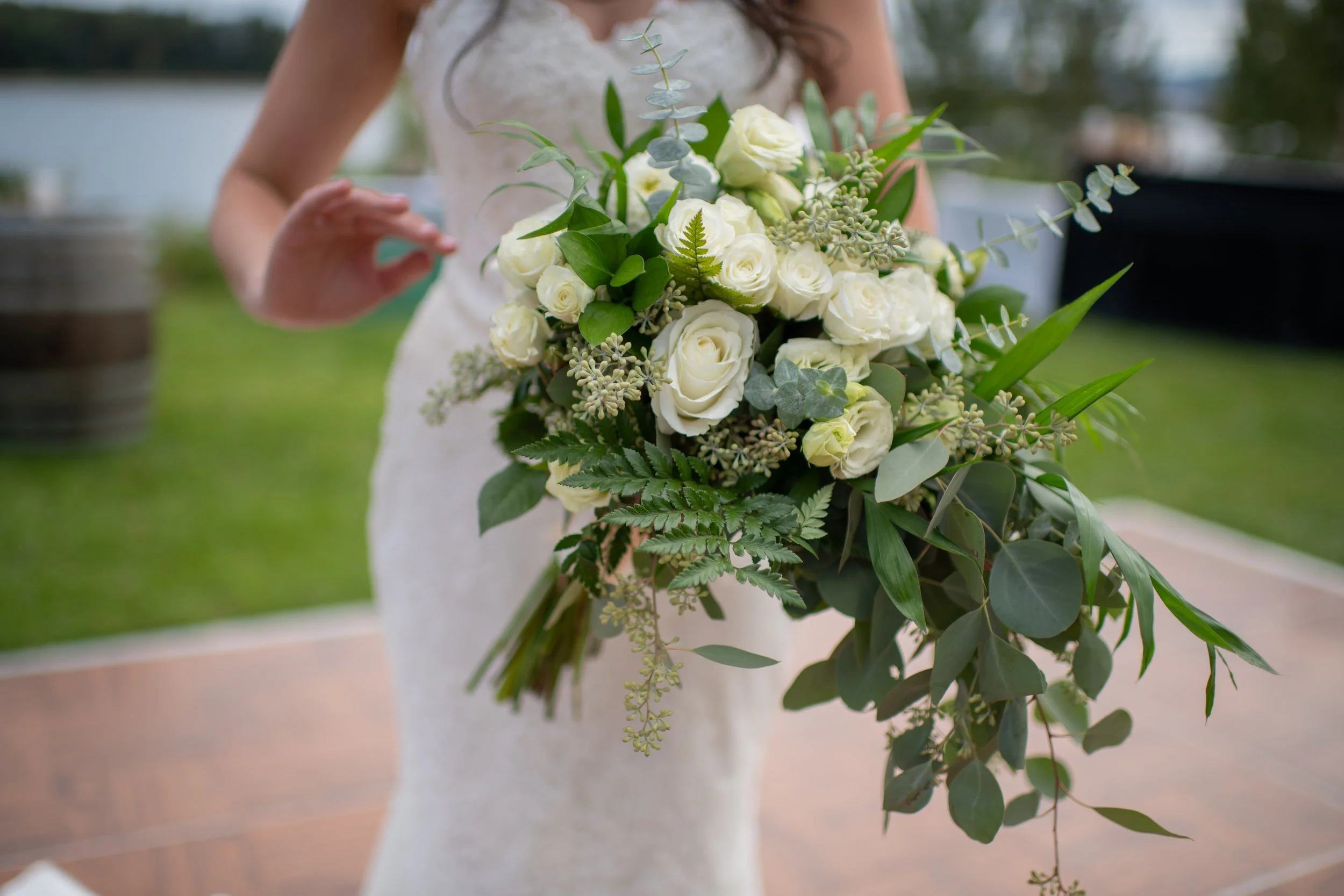 A bride holding a bouquet of white roses, green leaves, and various foliage outdoors near a body of water.