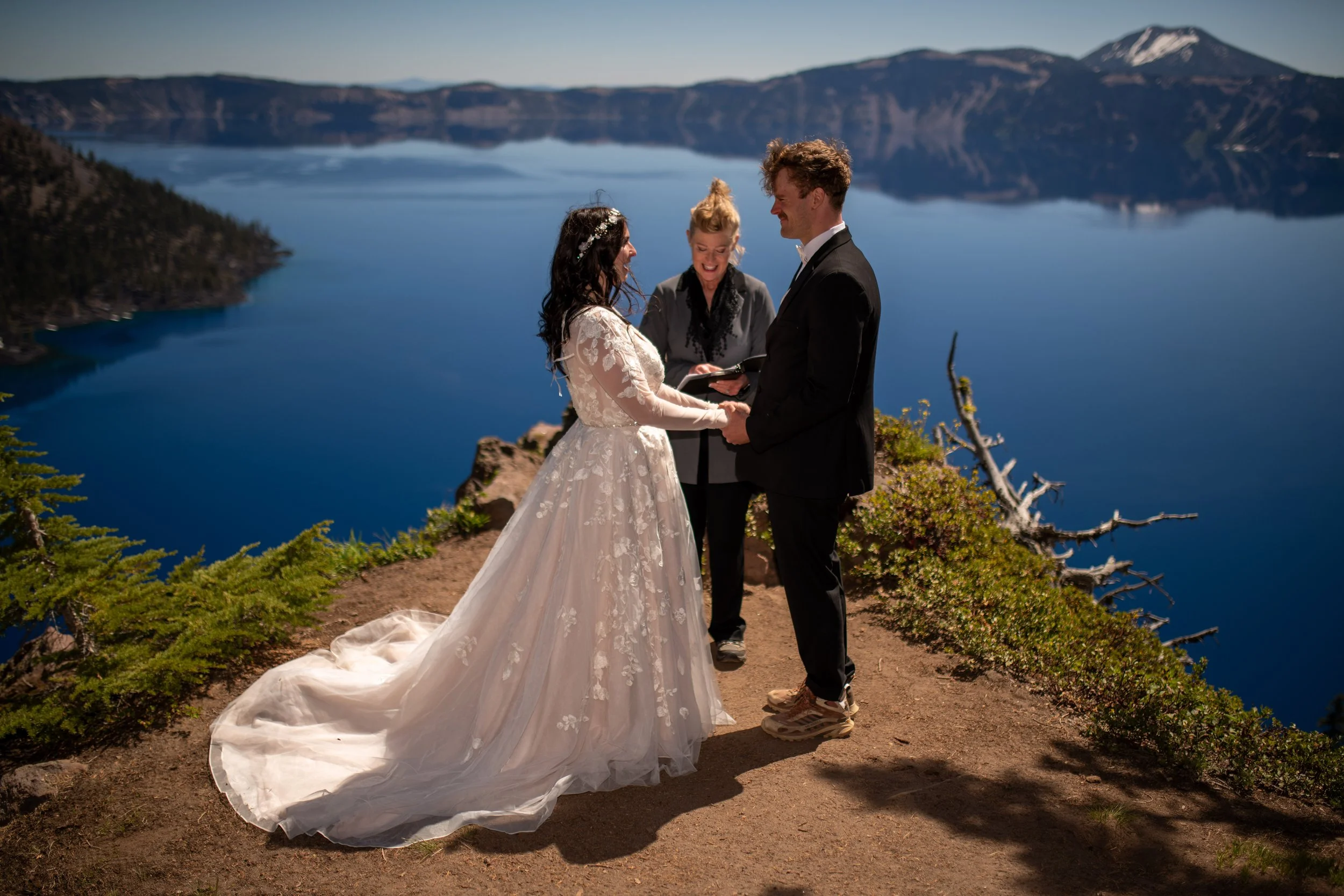 A couple getting married outdoors near a lake, with a woman officiant in the background, surrounded by mountains and clear sky.