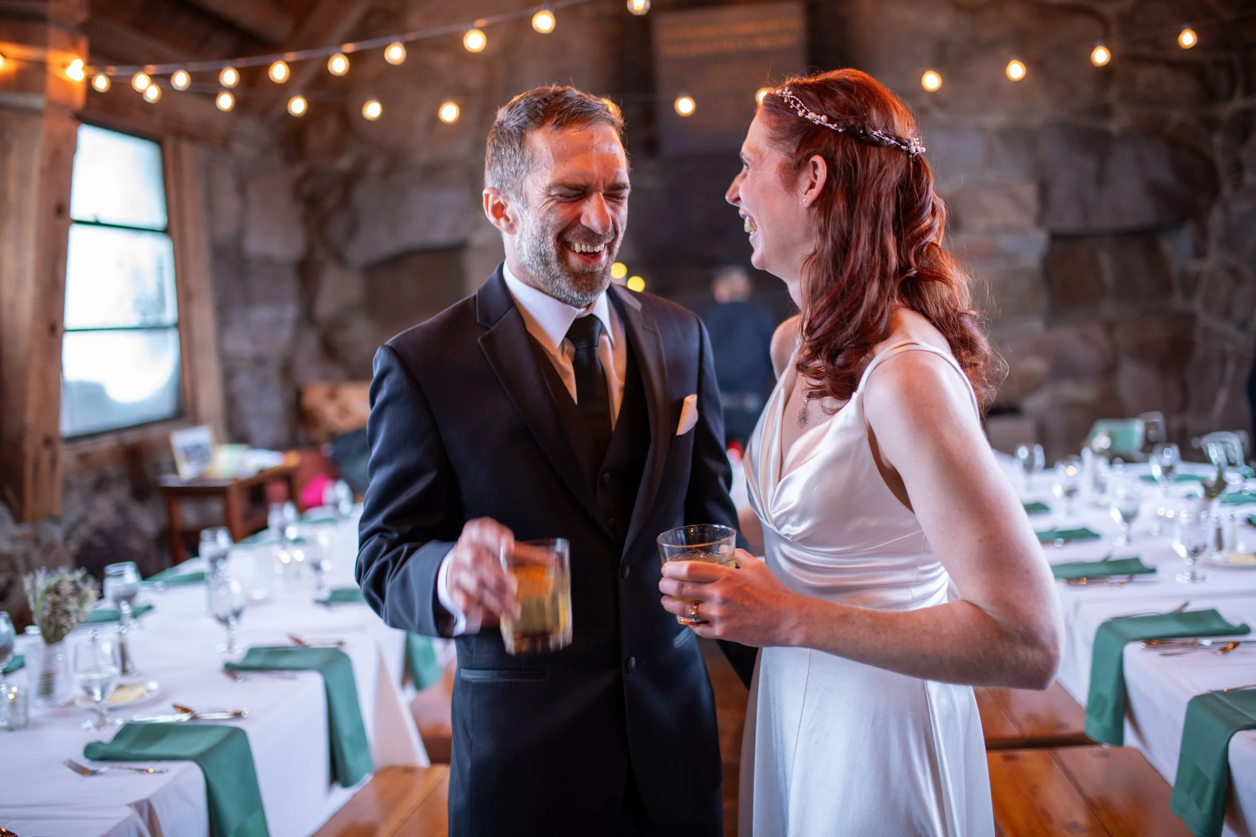 A smiling man and woman sharing a toast at a wedding reception, standing inside a rustic venue decorated with string lights, tables set for dinner, with wood walls and a window in the background.