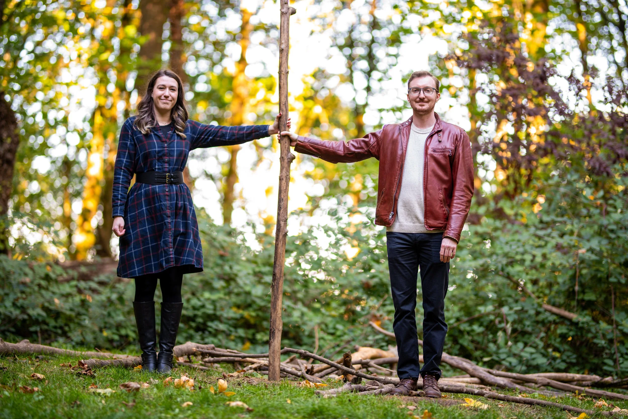A woman and a man are outdoors in a wooded area, holding a tree trunk upright between them. The woman is wearing a plaid dress and black boots, and the man is dressed in a leather jacket, light shirt, and glasses. They are standing on the ground with