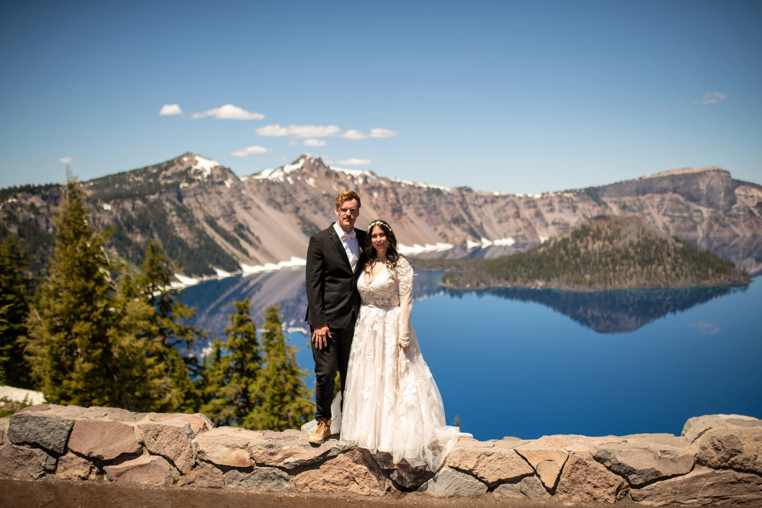 A couple dressed in wedding attire standing on a stone wall with a lake, mountains with snow patches, and a clear blue sky in the background.
