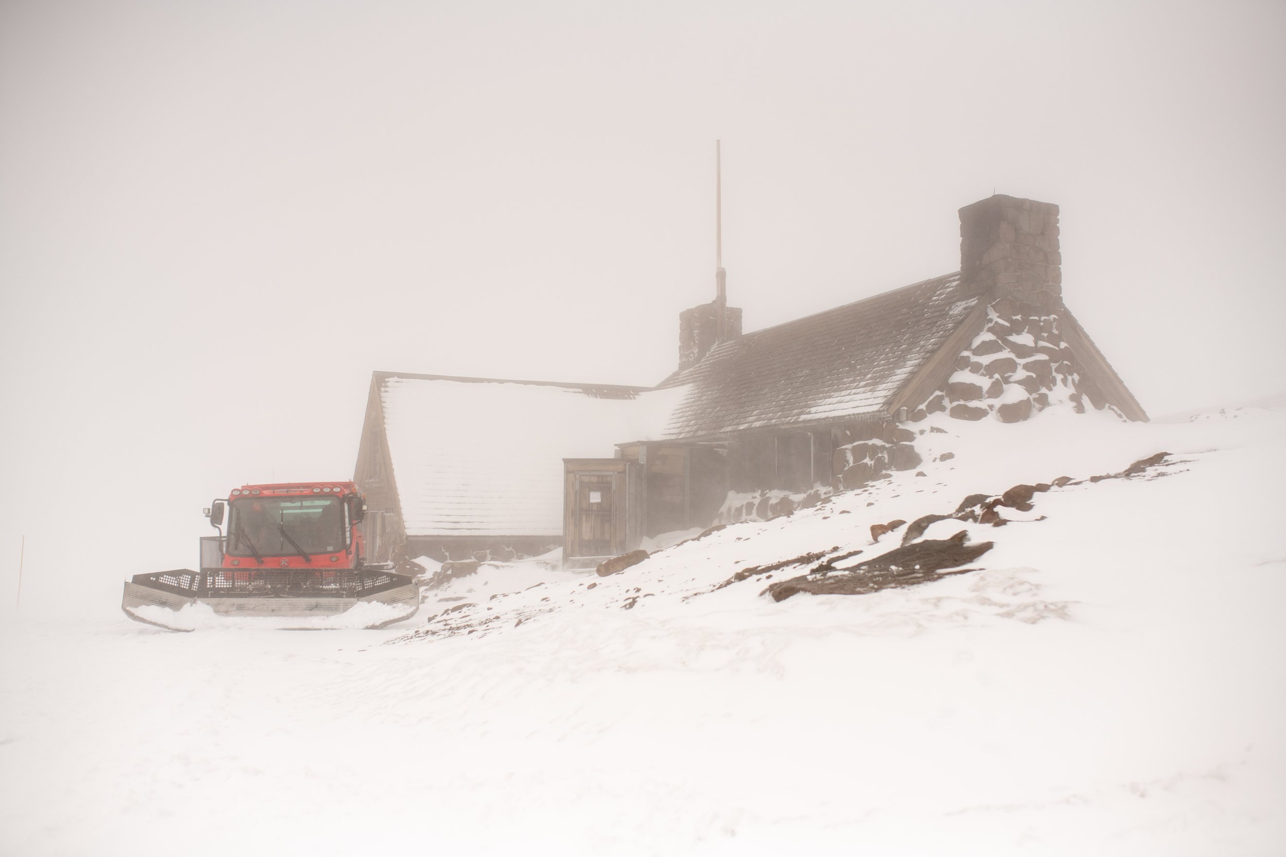 Snow-covered landscape with a rustic wooden house, a brick chimney, and a snowplow truck parked nearby, all shrouded in fog.