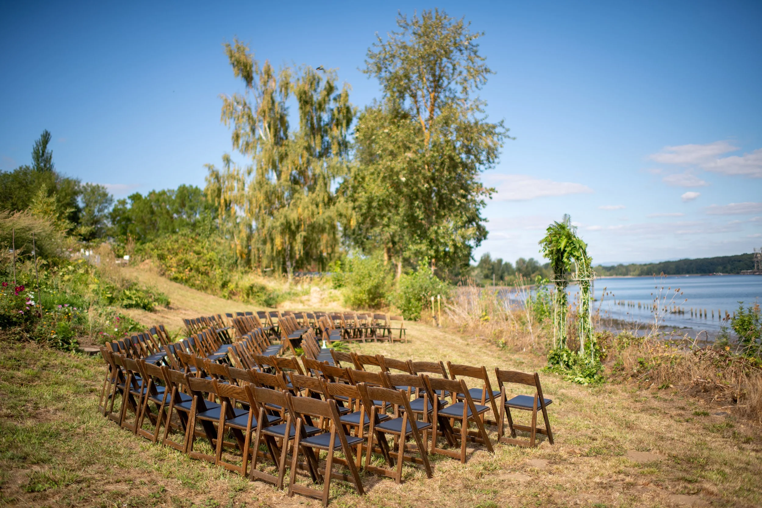 Rows of wooden chairs arranged outdoors near a lake with trees and a clear blue sky in the background.