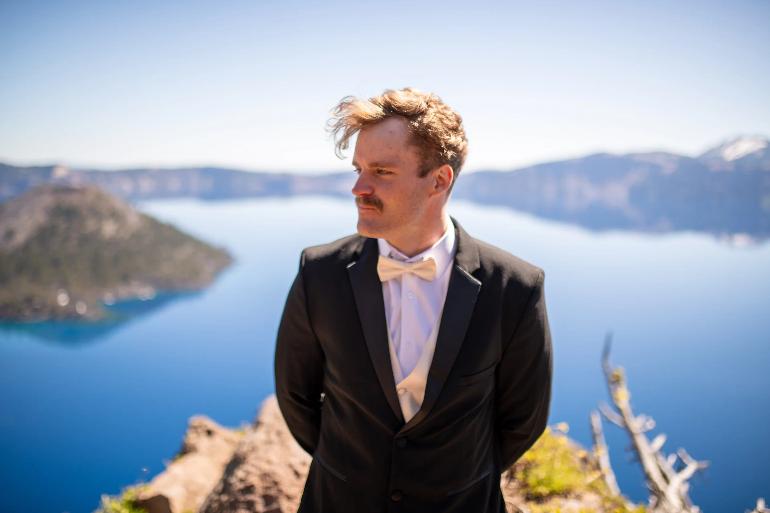 A man in a tuxedo with a white bow tie standing outdoors near a body of water and mountains in the background.