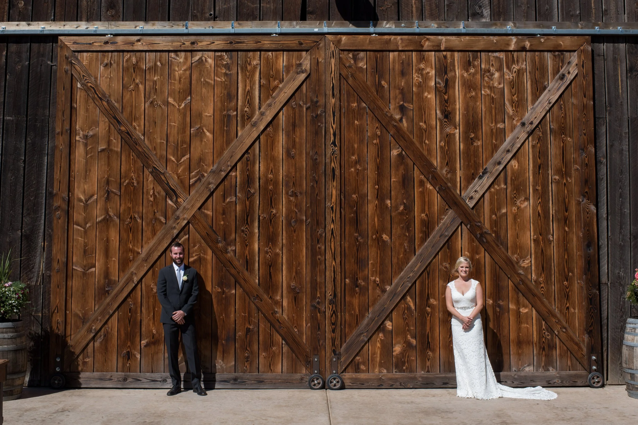 A bride and groom are standing in front of a large wooden sliding barn door at a wedding. The groom is wearing a dark suit with a white shirt and bow tie, and the bride is in a white wedding dress. The setting appears to be outdoors.
