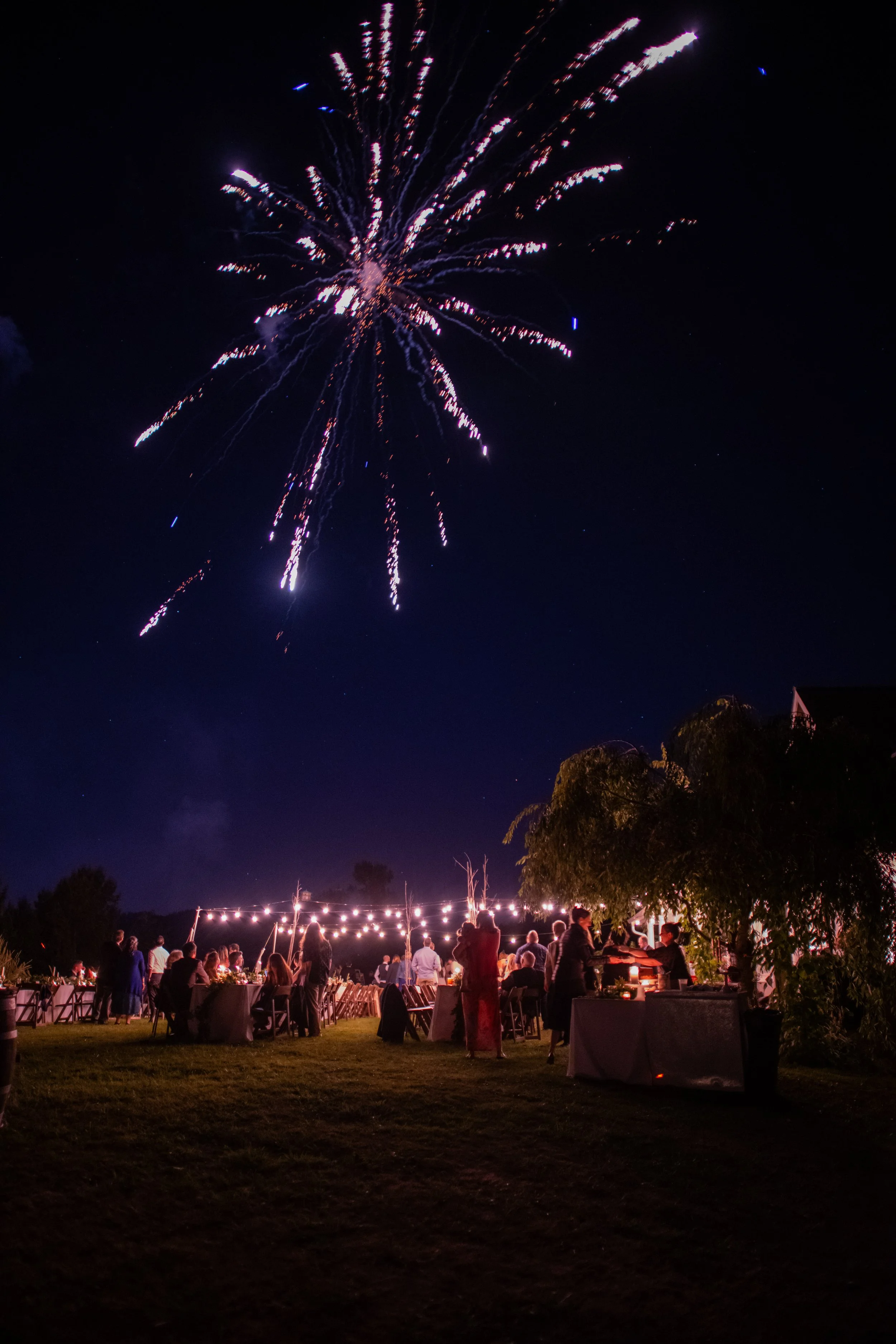 Night scene of a celebration with fireworks in the sky, people gathered around tables with string lights, and trees and houses in the background.