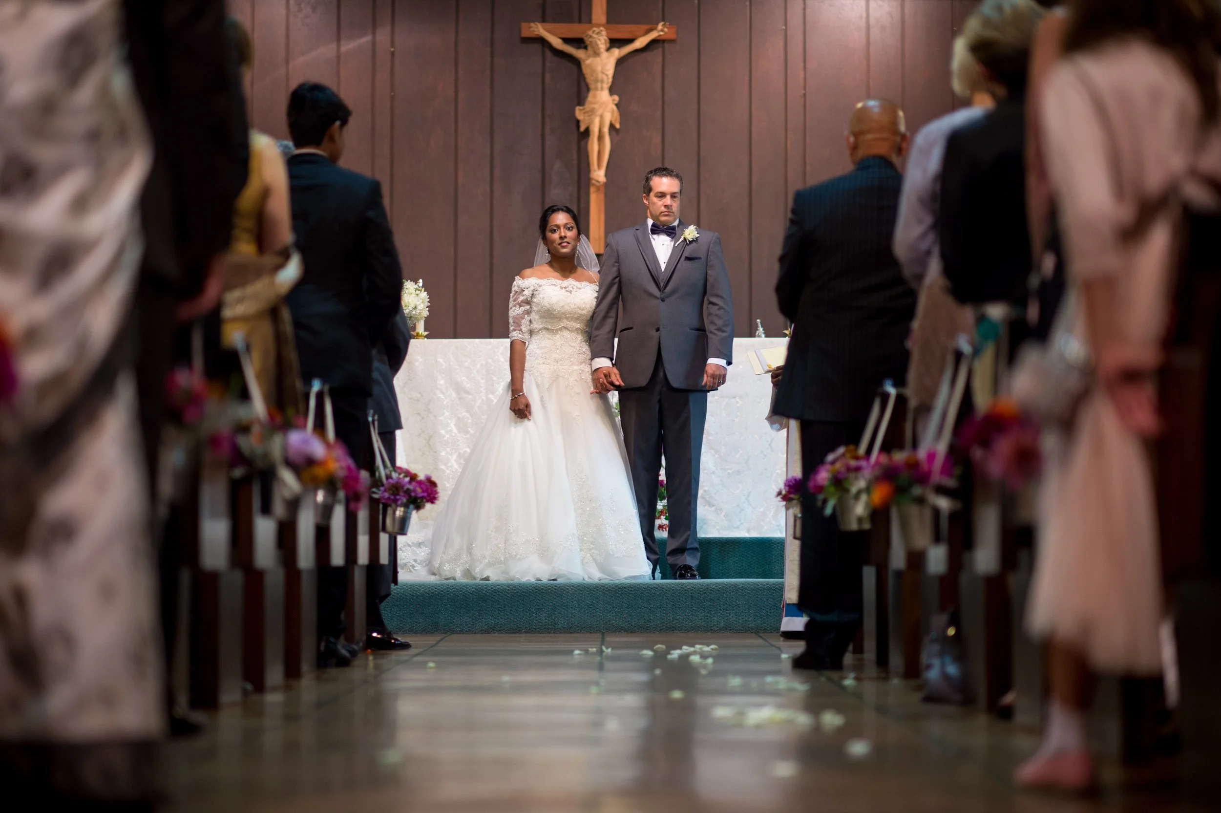 A bride and groom standing together at the altar during their wedding ceremony inside a church, with guests seated in pews on either side, facing the couple, and a crucifix hanging on the wall behind them.