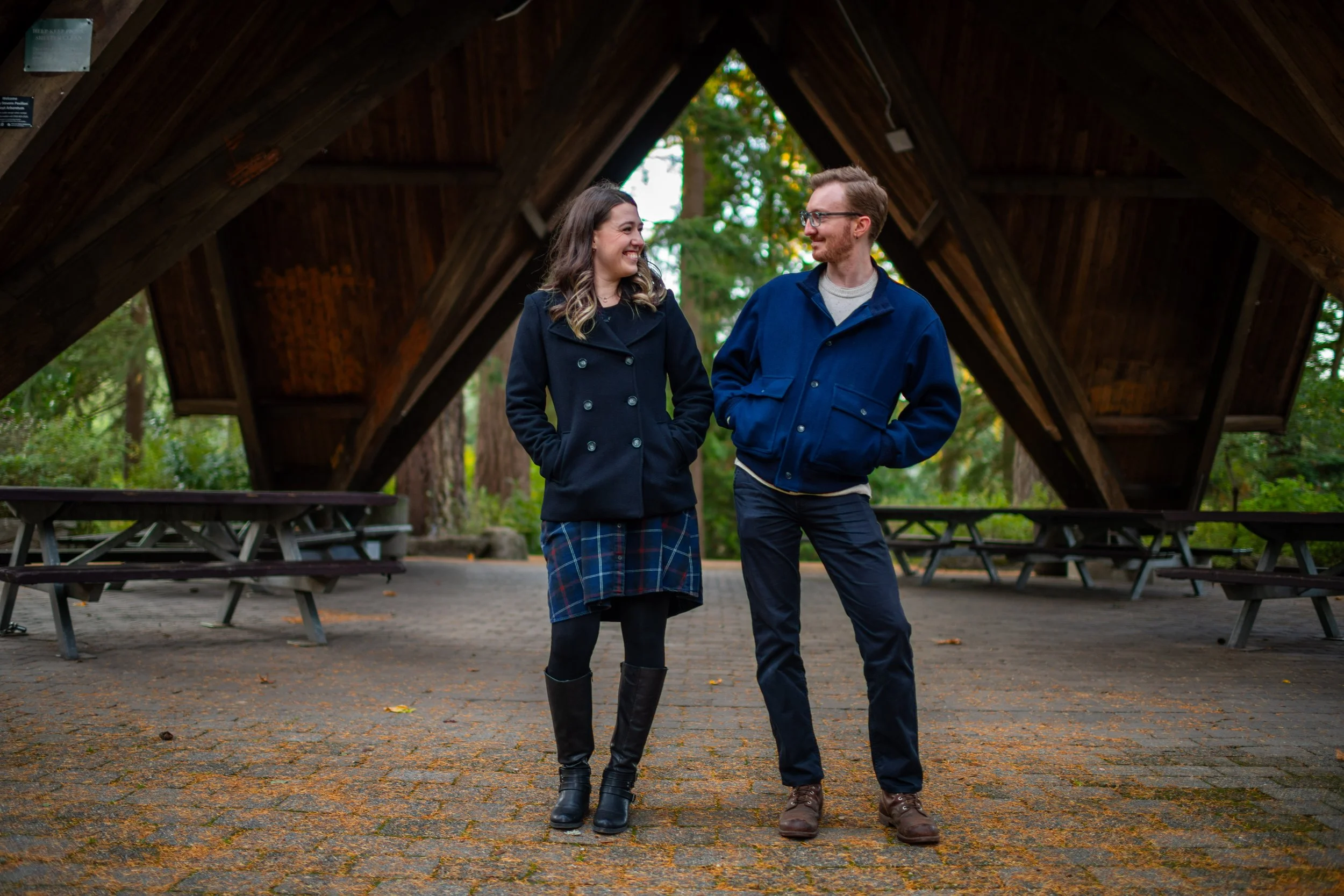 A woman and man walking and smiling at each other in a park under a wooden pavilion with picnic tables in the background.