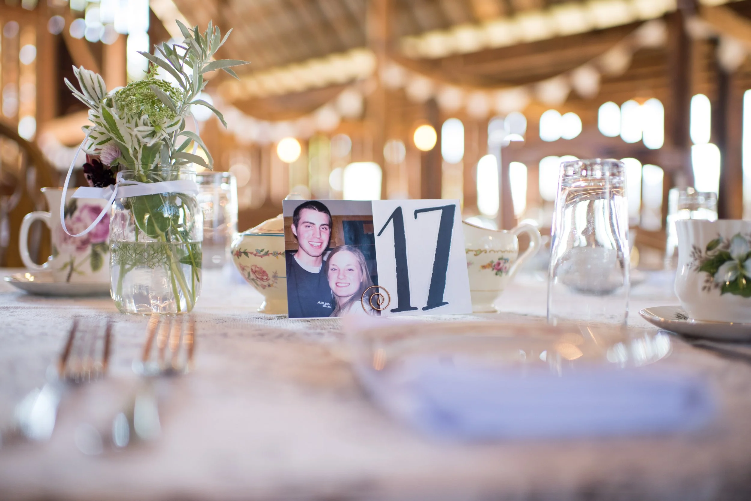 Table setting with a floral centerpiece in a glass jar, a framed photograph of a young man and woman, a numbered table card, teapot, glass of water, and china cups in a rustic venue with warm lighting and wooden beams.