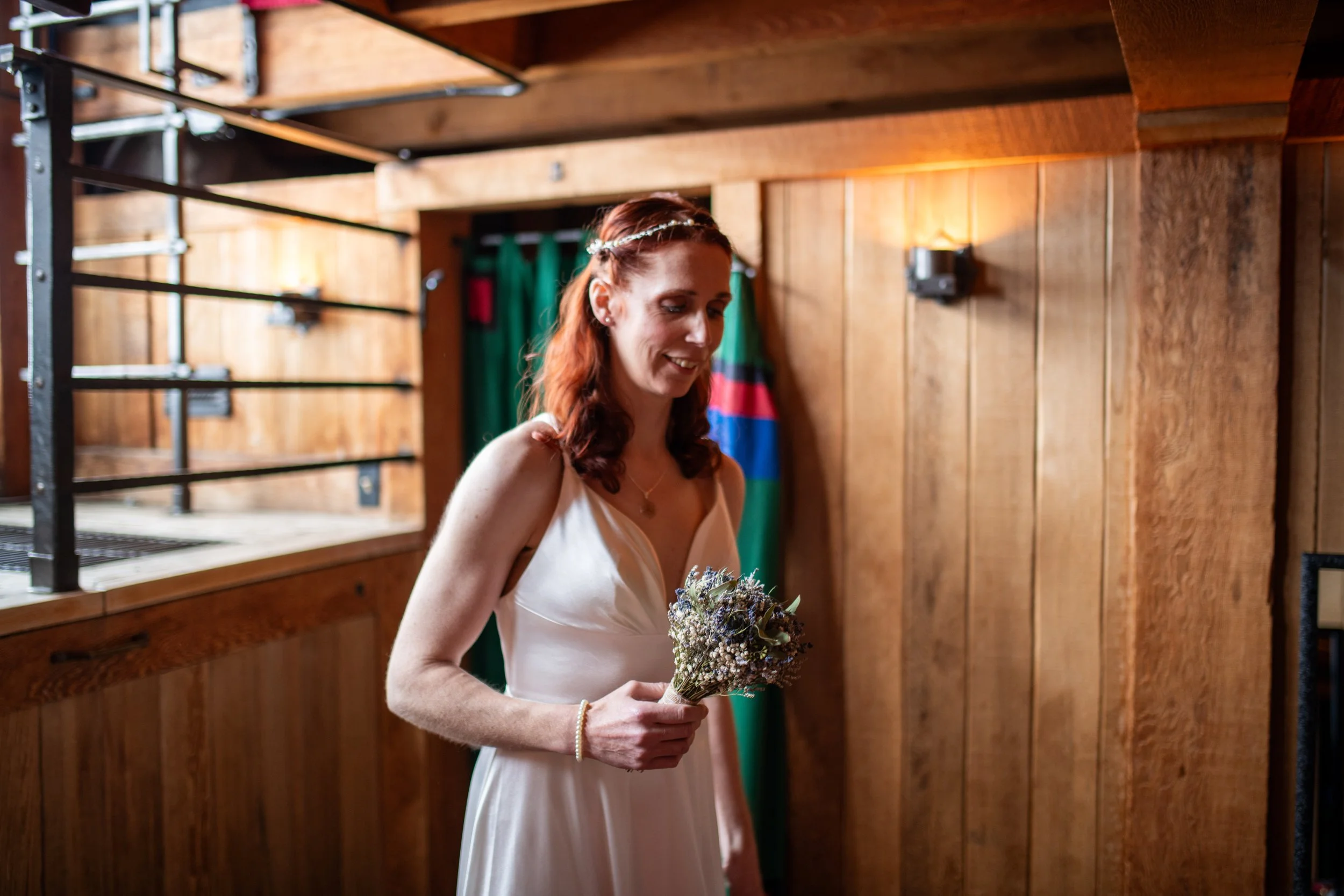 A woman with red hair, wearing a white dress and a floral headband, holding a bouquet of flowers, standing indoors with wooden panel walls and green and red fabric in the background.