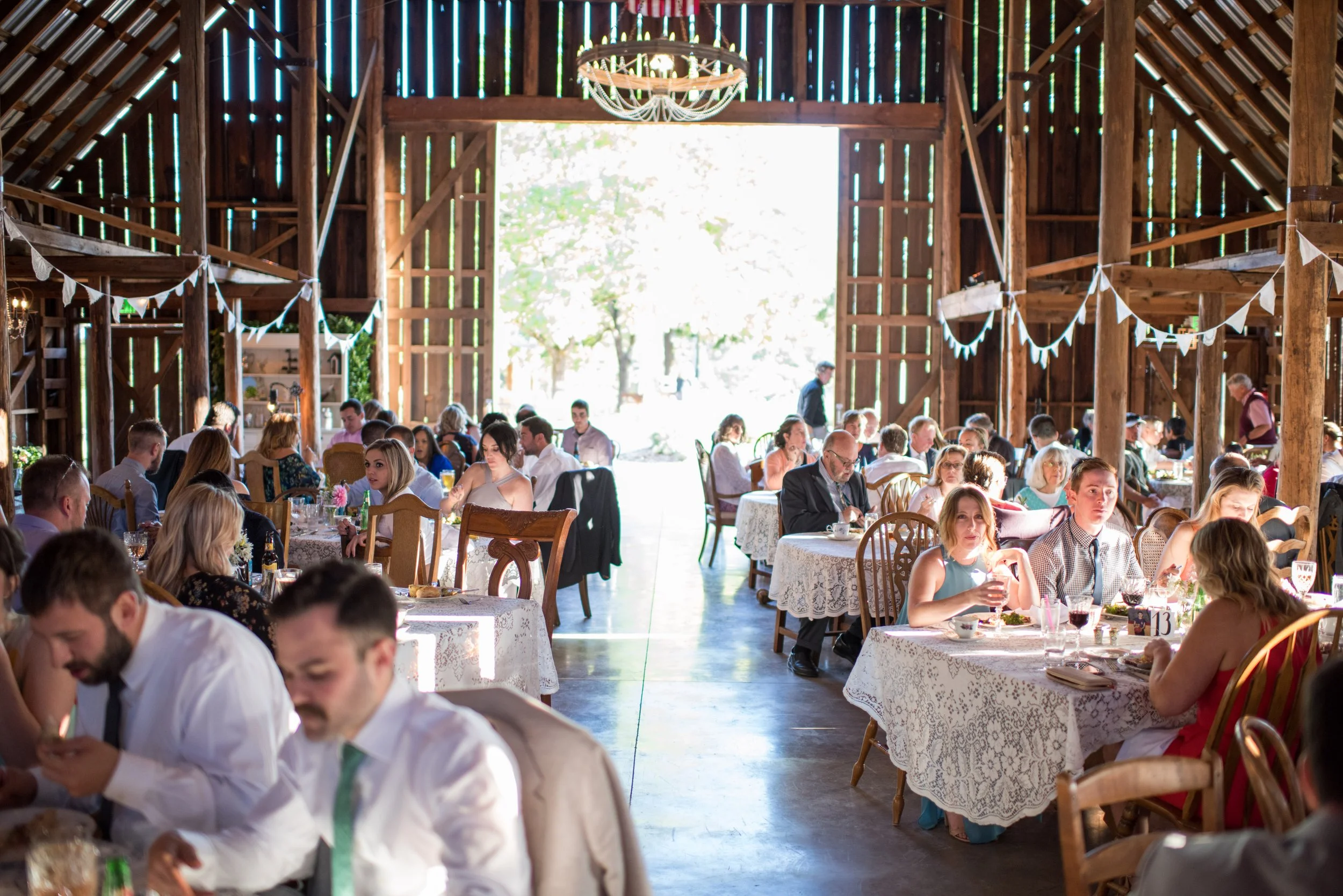 Inside a rustic barn decorated for a celebration, with people sitting at tables enjoying a meal.