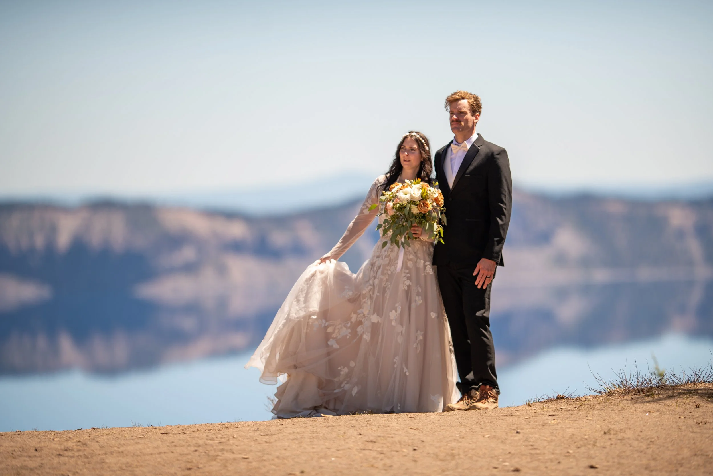 A bride and groom standing outdoors on a hilltop with a view of mountains and sky in the background, dressed in wedding attire.