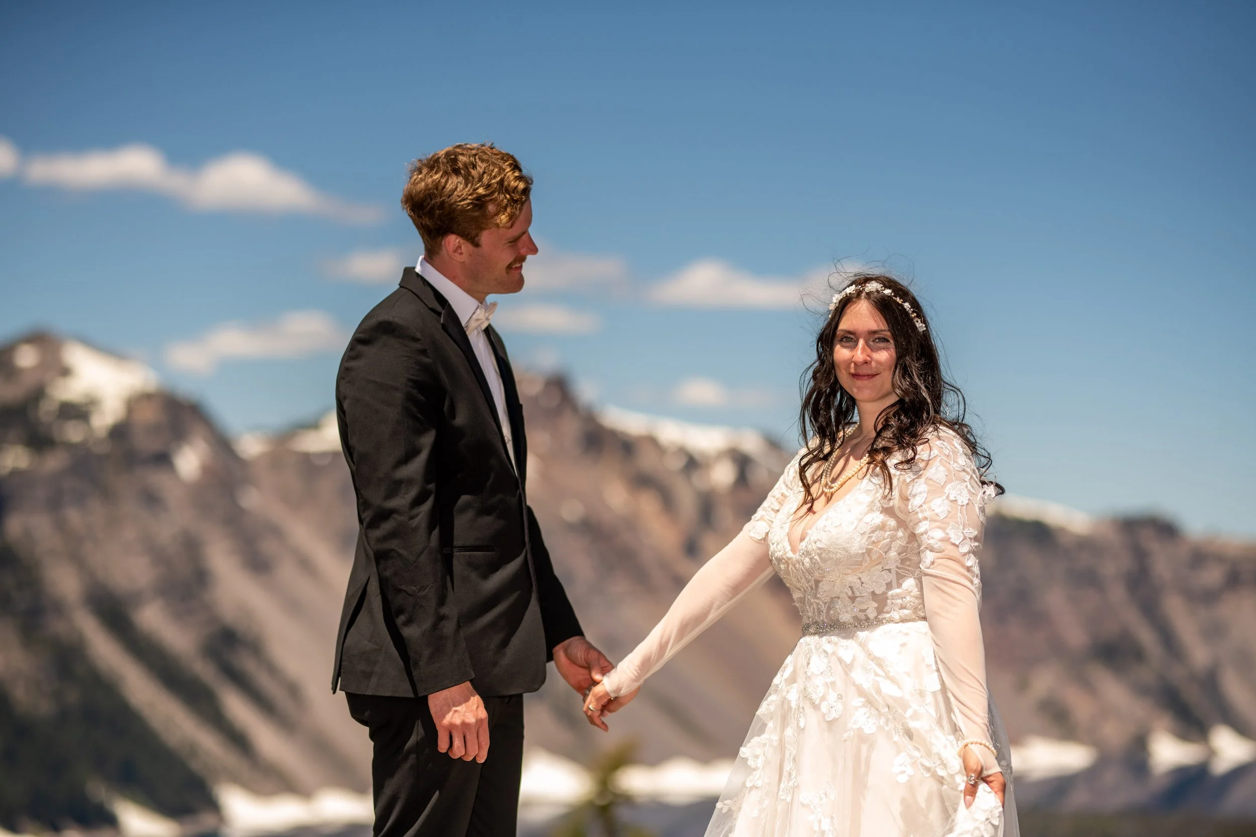 A couple dressed in wedding attire holding hands outdoors against a mountainous landscape under a blue sky with white clouds.