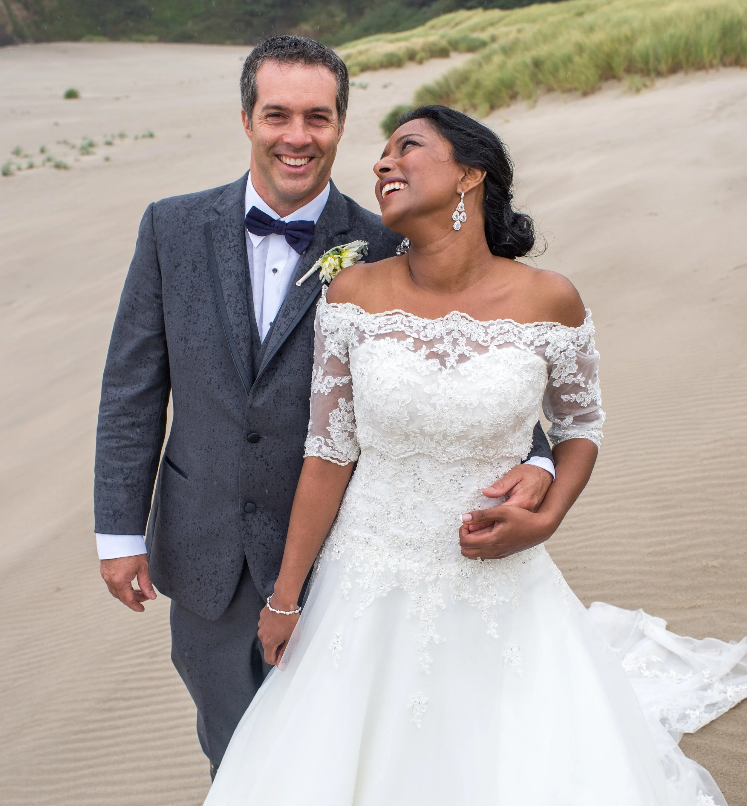 A smiling couple, a man in a tuxedo and a woman in a white lace wedding dress, standing close together on a sandy beach with sand dunes and green Grass in the background, holding hands.