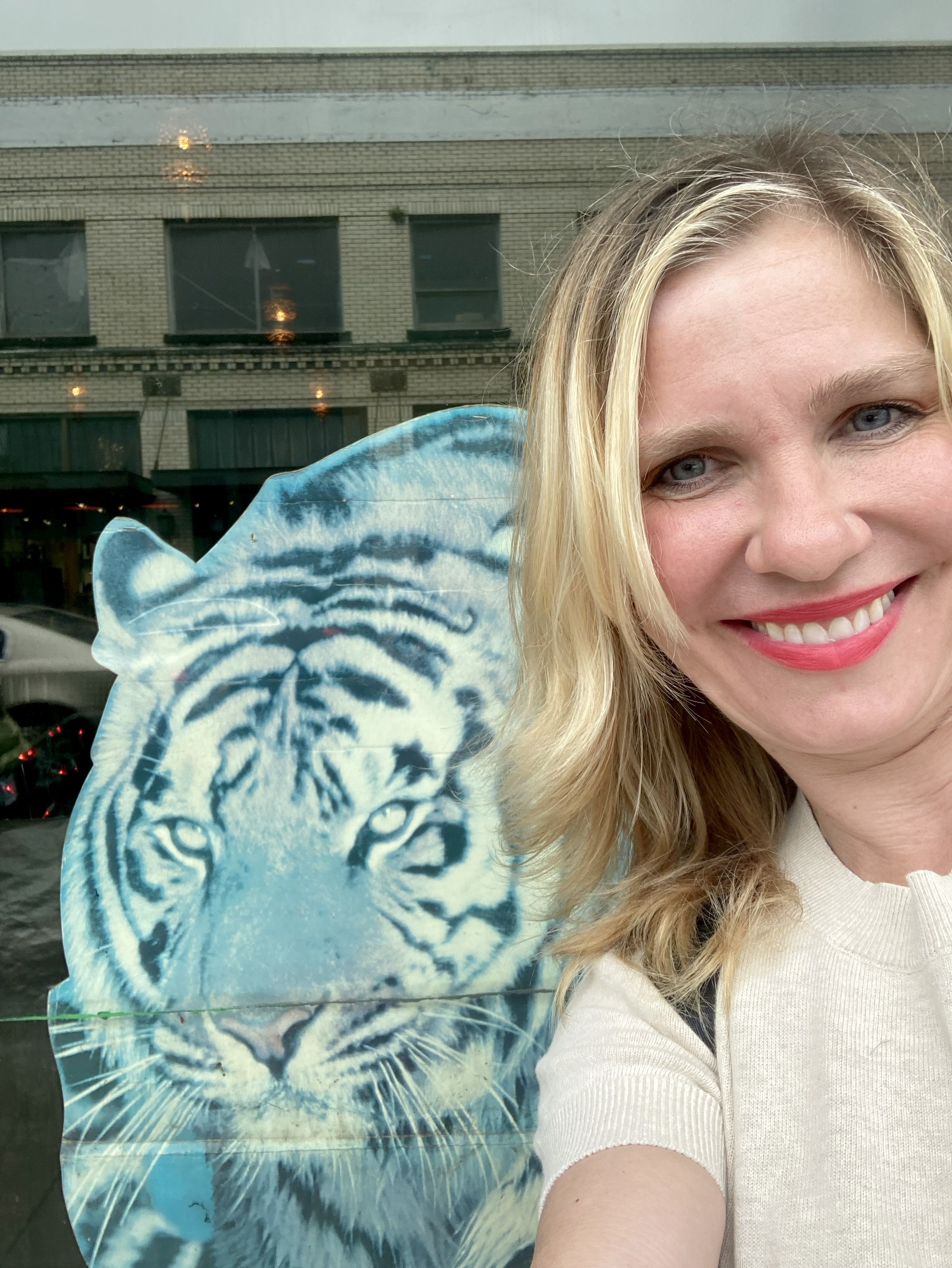 A woman with blonde hair and blue eyes smiling, taking a selfie in front of a building with large windows. She is wearing a light-colored top and holding a blue tiger-themed umbrella.