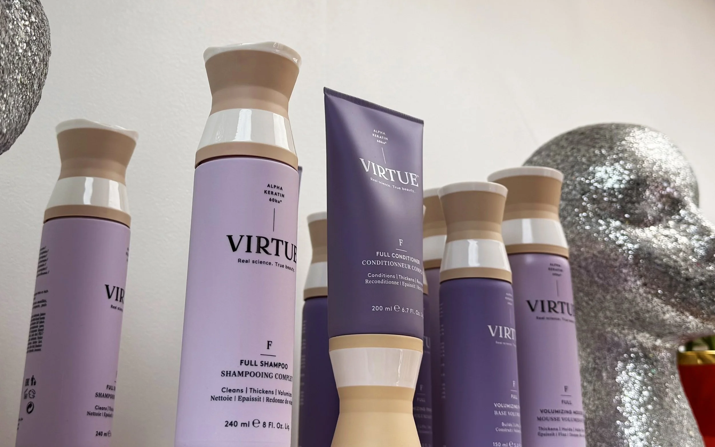 Multiple bottles of Virtue shampoo and conditioner, purple with beige caps, displayed on a white surface indoors with decorative silver balls in the background.