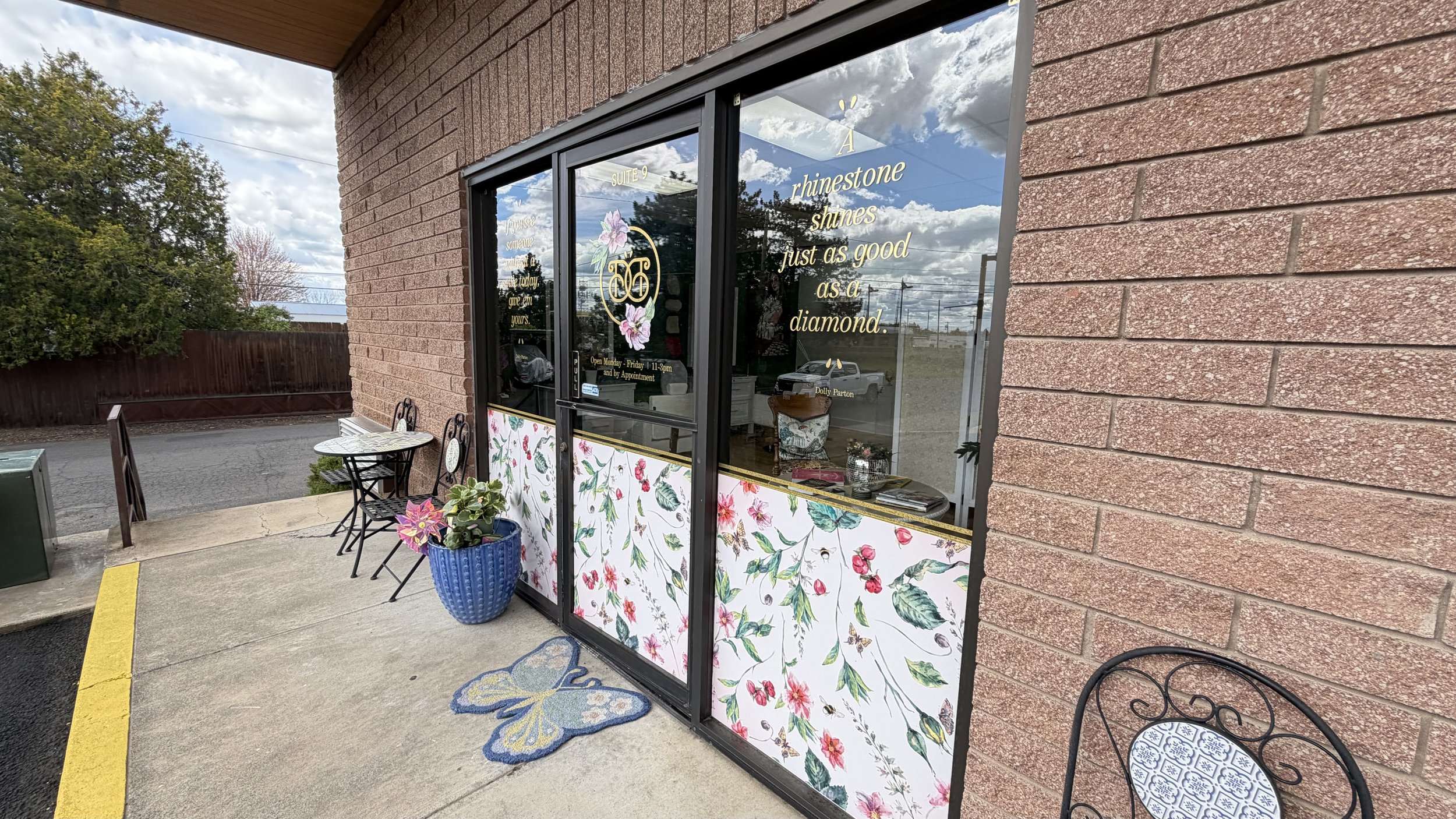 Storefront with large glass doors decorated with floral patterns and gold lettering, featuring signs about rhinestone shines and jewelry, with outdoor seating including two black metal chairs and a blue potted plant outside.