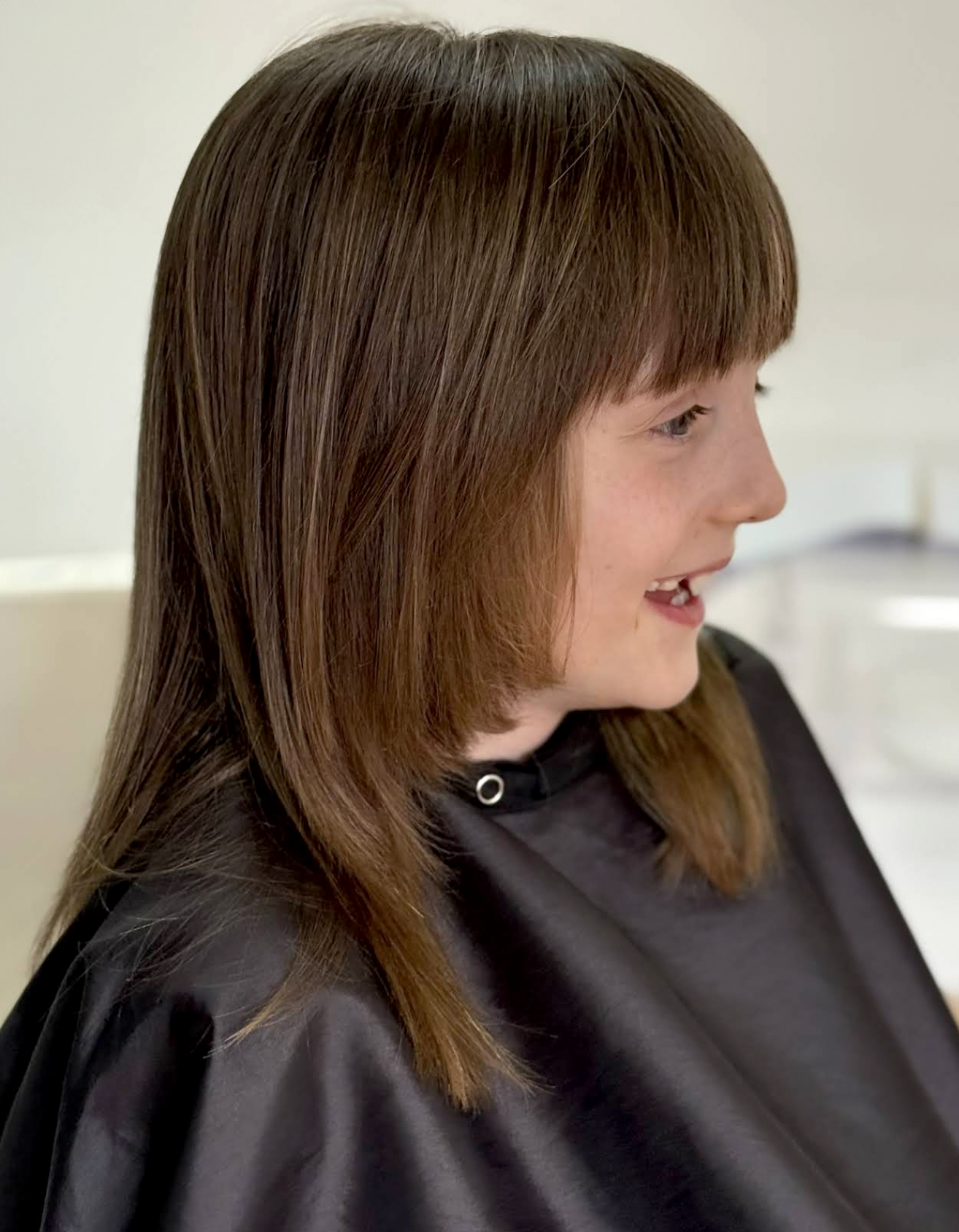 Side profile of a woman with reddish-brown hair, wearing a black salon cape during a haircut in a salon.
