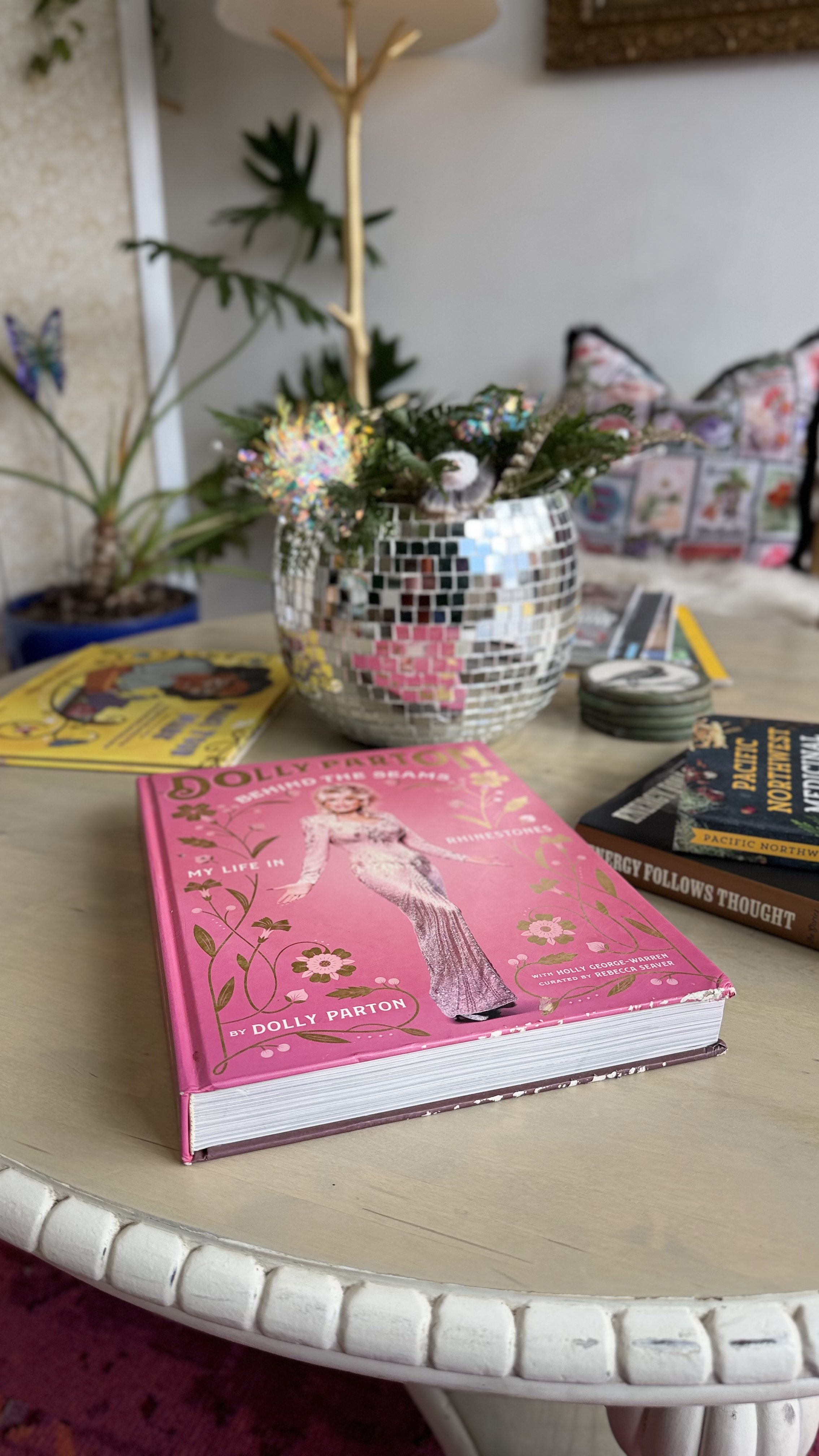 A pink book titled "Dolly Parton" is on a white round table, along with a mosaic vase with some greenery, and other books. In the background, there are plants and a decorative butterfly.