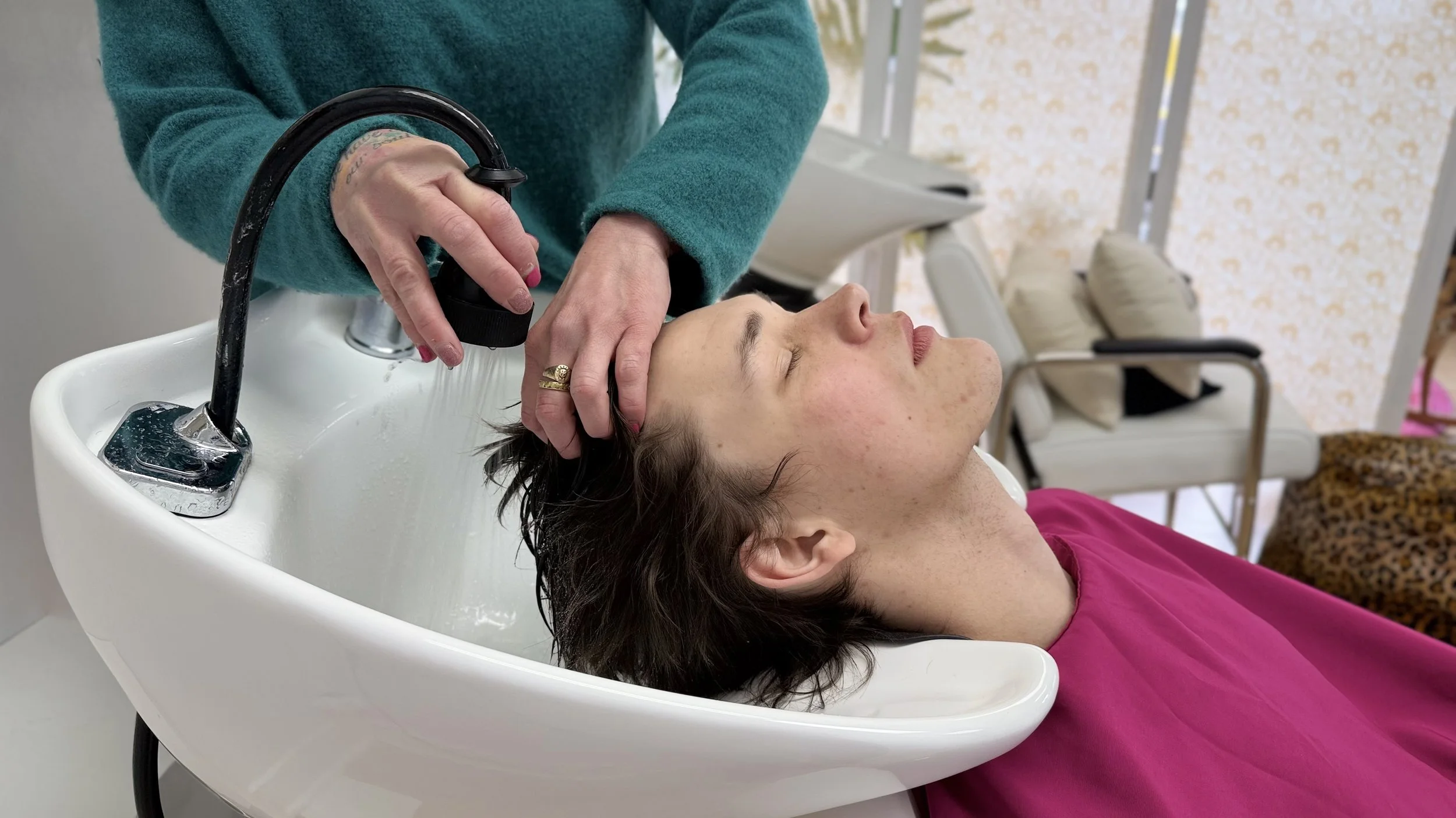 A woman with short dark hair lying back at a salon sink with her eyes closed as a stylist washes her hair. The woman is wearing a pink top.