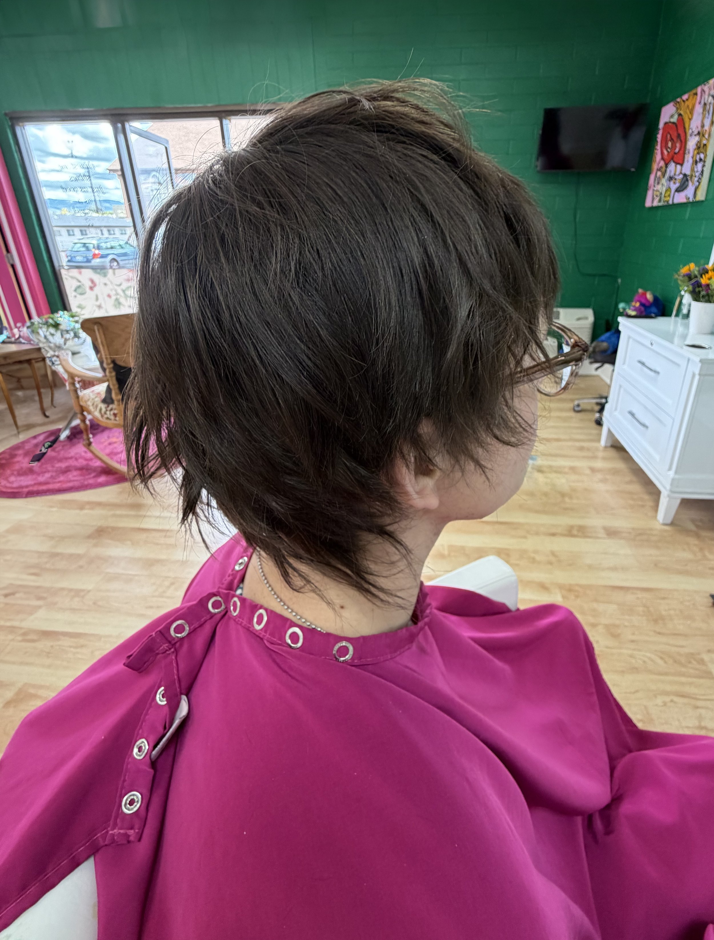A woman with short brown hair wearing a pink salon cape, standing inside a hair salon with green walls, a TV on the wall, and a painting beside it.