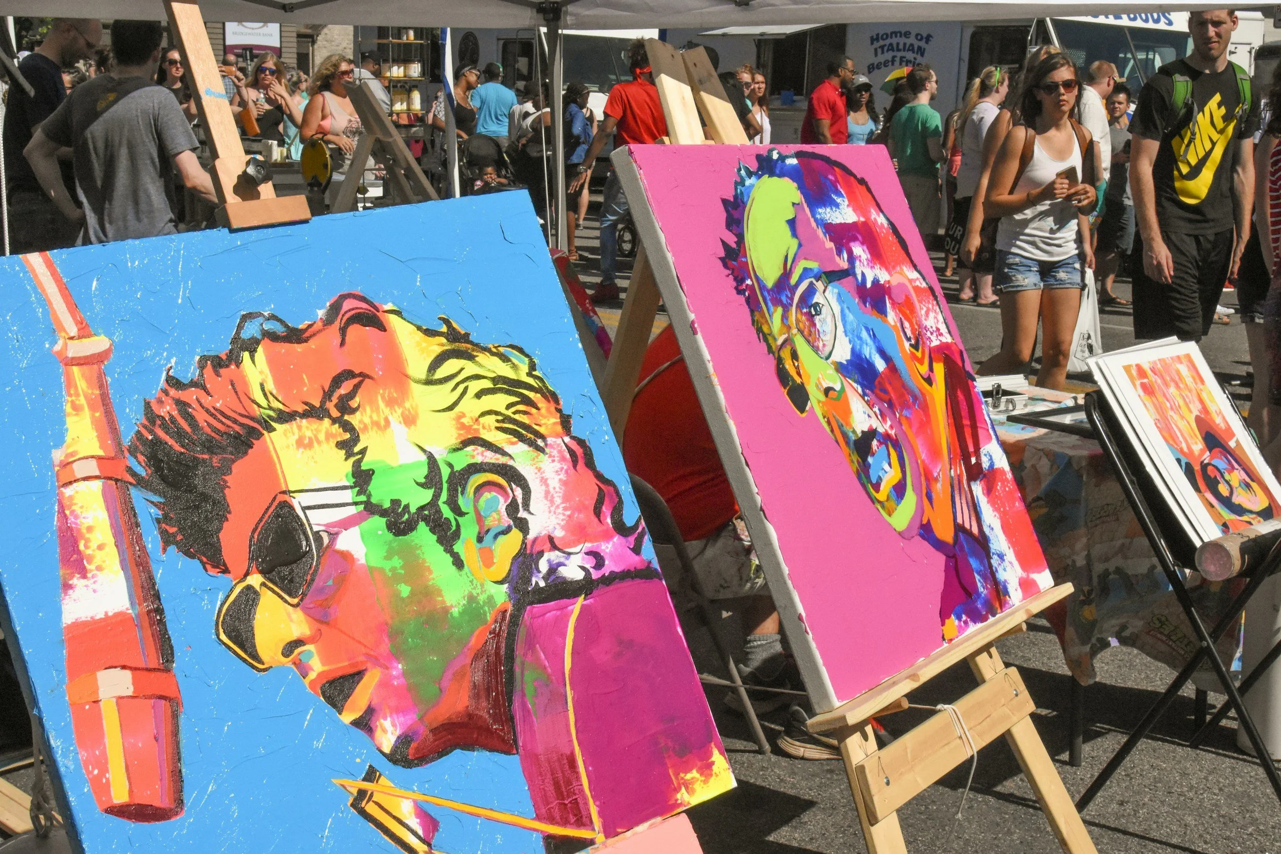 Multiple colorful paintings, including a pop art style portrait of a person with orange and yellow hues and a background with bright pink and purple, displayed on easels at an outdoor art market with people walking by.