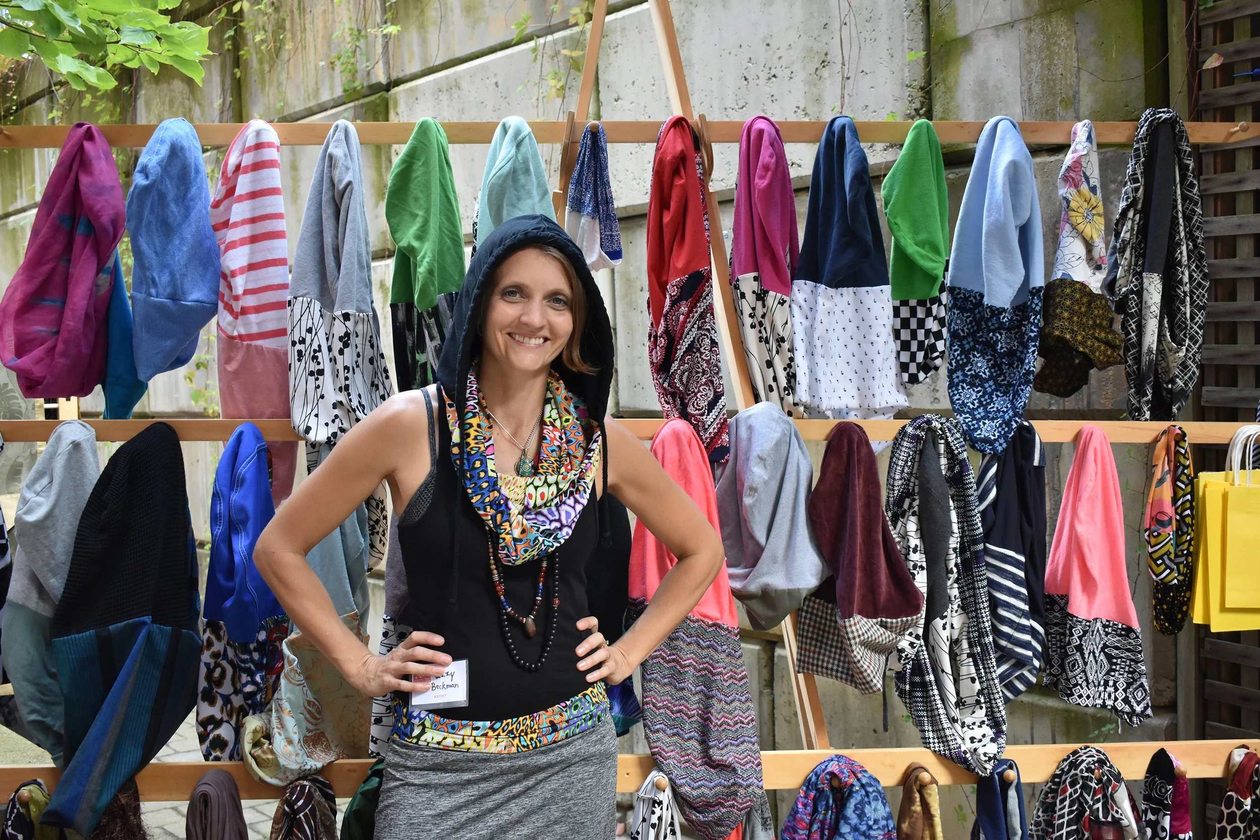 A woman smiling at a clothing and accessories display outdoors with colorful scarves hanging on a wooden rack