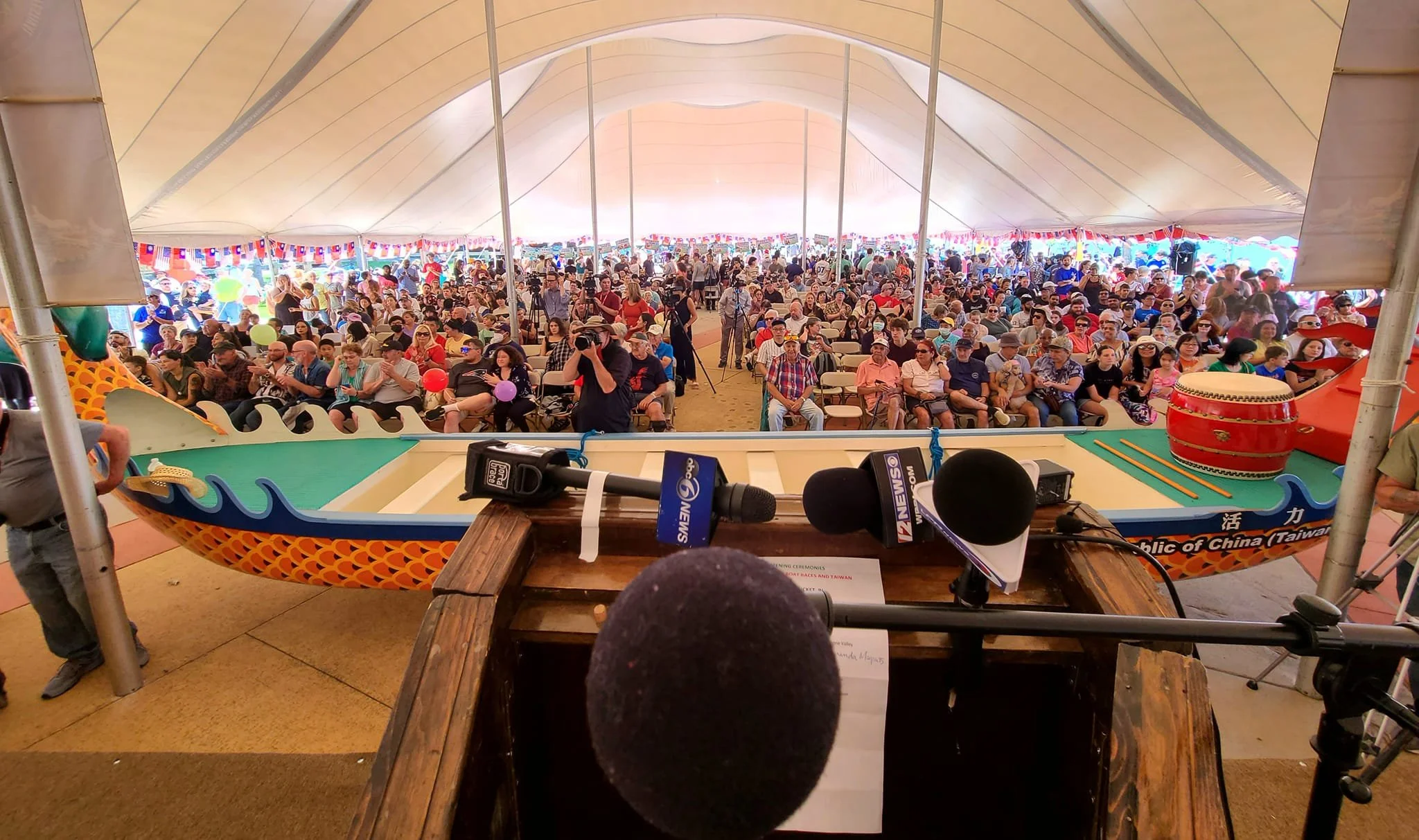 Crowd gathered under a large tent at an outdoor event, with a game table in the foreground, surrounded by microphones and news cameras.