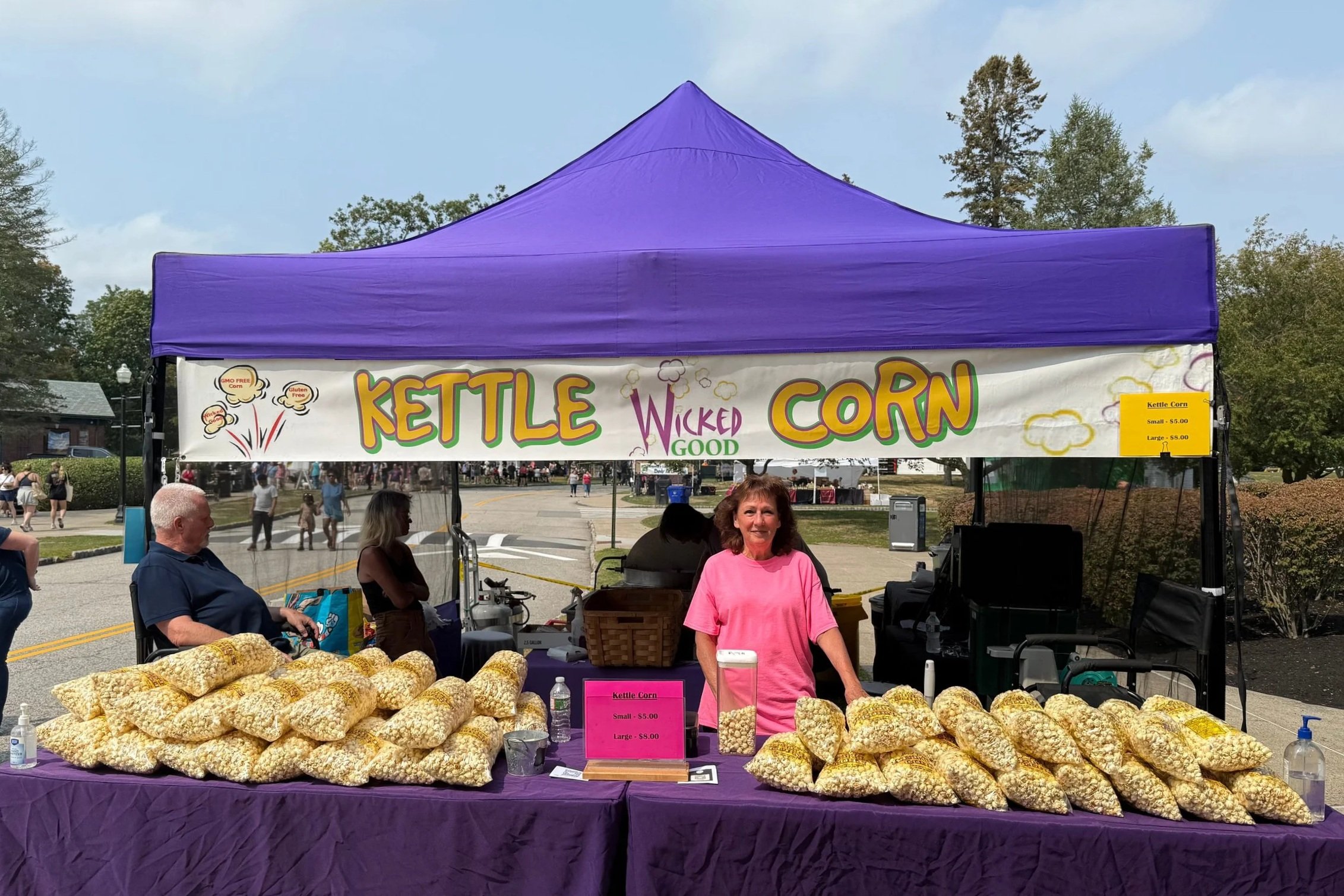 At a fair, a vendor's booth has a purple tent with a sign that reads 'Kettle Wicked Good Corn,' offering small and large portions for sale at $5.00 and $8.00. The table is filled with bags of popcorn. Several people are seen in front of and around the booth, with trees and a street in the background.