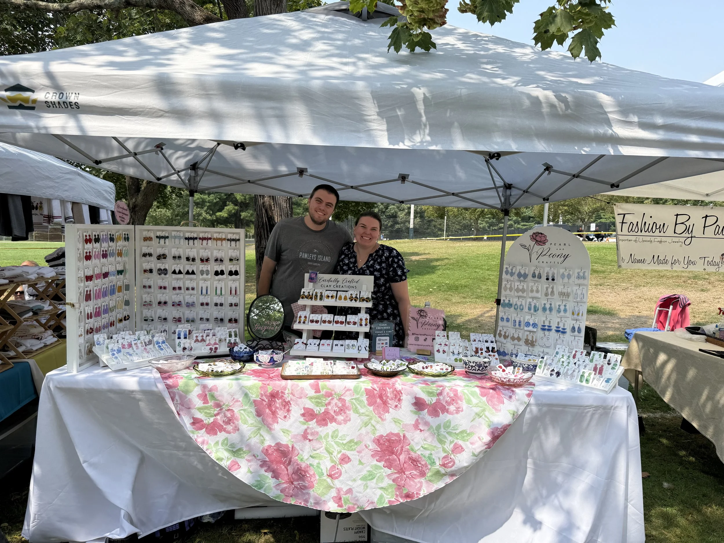 View of an outdoor craft fair booth under a white canopy tent, displaying various jewelry items on white display boards and tables with pink, green, and white floral tablecloths. Two people, a man and a woman, stand behind the display, smiling at the camera. There are trees and a grassy park in the background.