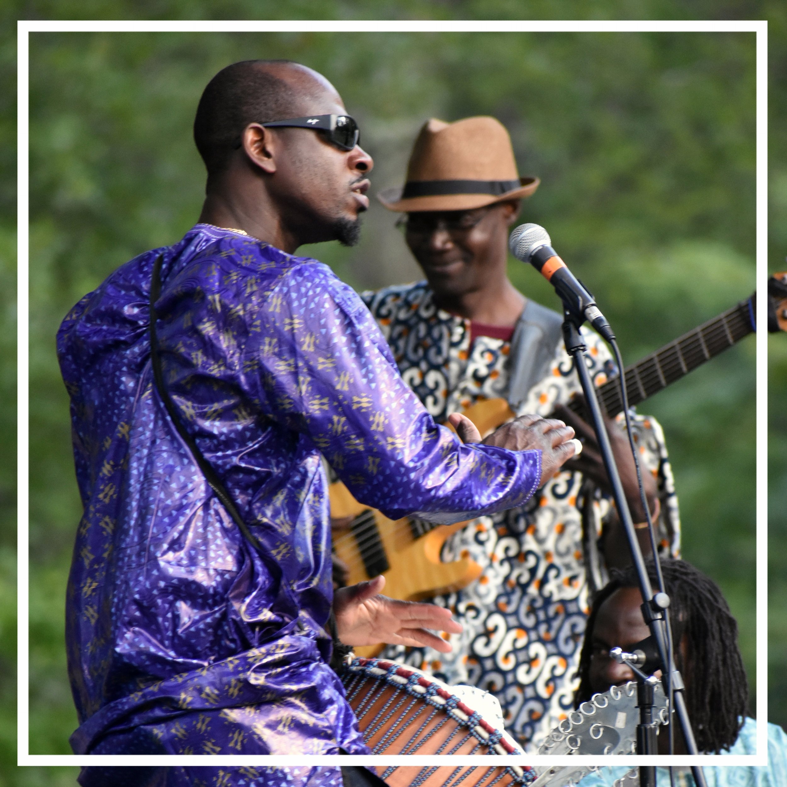 Two men performing music outdoors. The man in the foreground wears sunglasses and a blue patterned shirt, and the man in the background wears a brown hat and patterned shirt, playing a guitar and singing into a microphone.