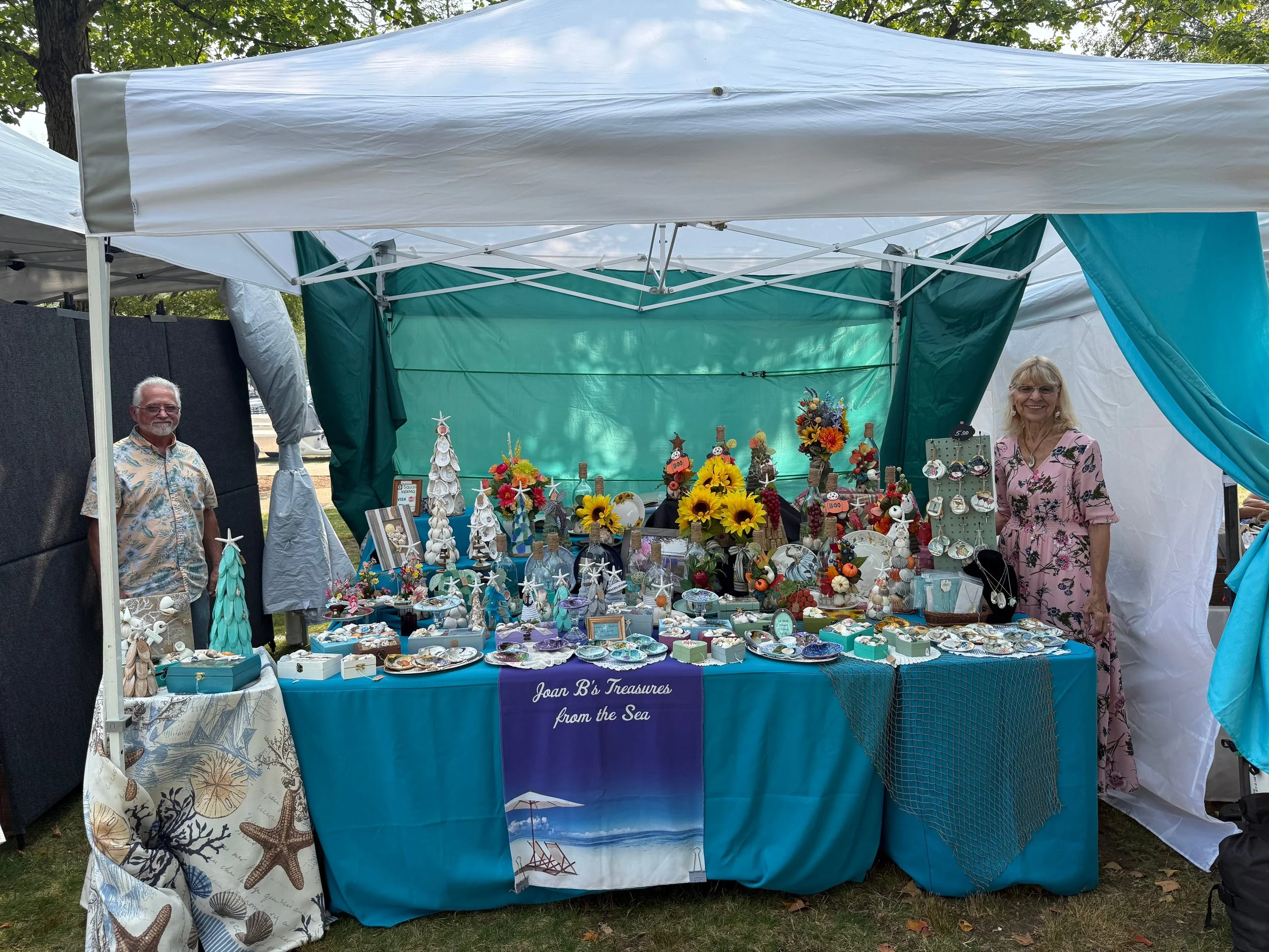 A woman and a man standing behind a table at an outdoor market stall displaying seashell, coastal-themed jewelry, and decorative items. The stall has a blue tablecloth and a sign that reads "Joan B's Treasures from the Sea." The background shows trees and sky, with the stall covered by a canopy.