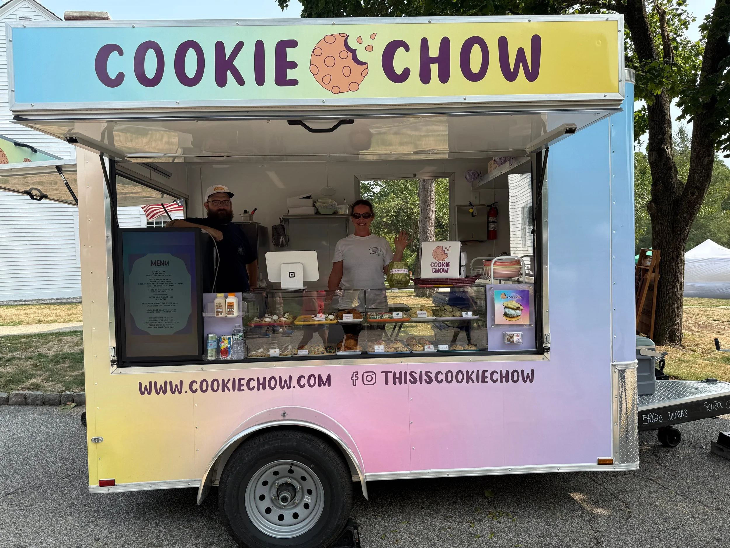 A colorful cookie food truck with the words 'Cookie Chow' on the sign, two people inside smiling, with various cookies on display in the front glass case.
