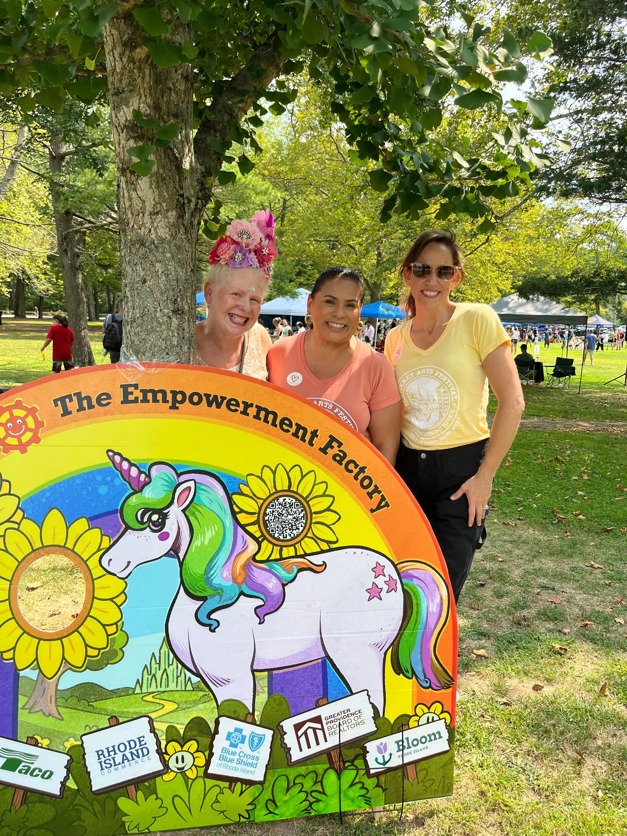 Three women are standing outdoors behind a colorful cutout board with a unicorn and sunflower design, smiling at the camera during a festival or event in a park with tents and trees in the background.