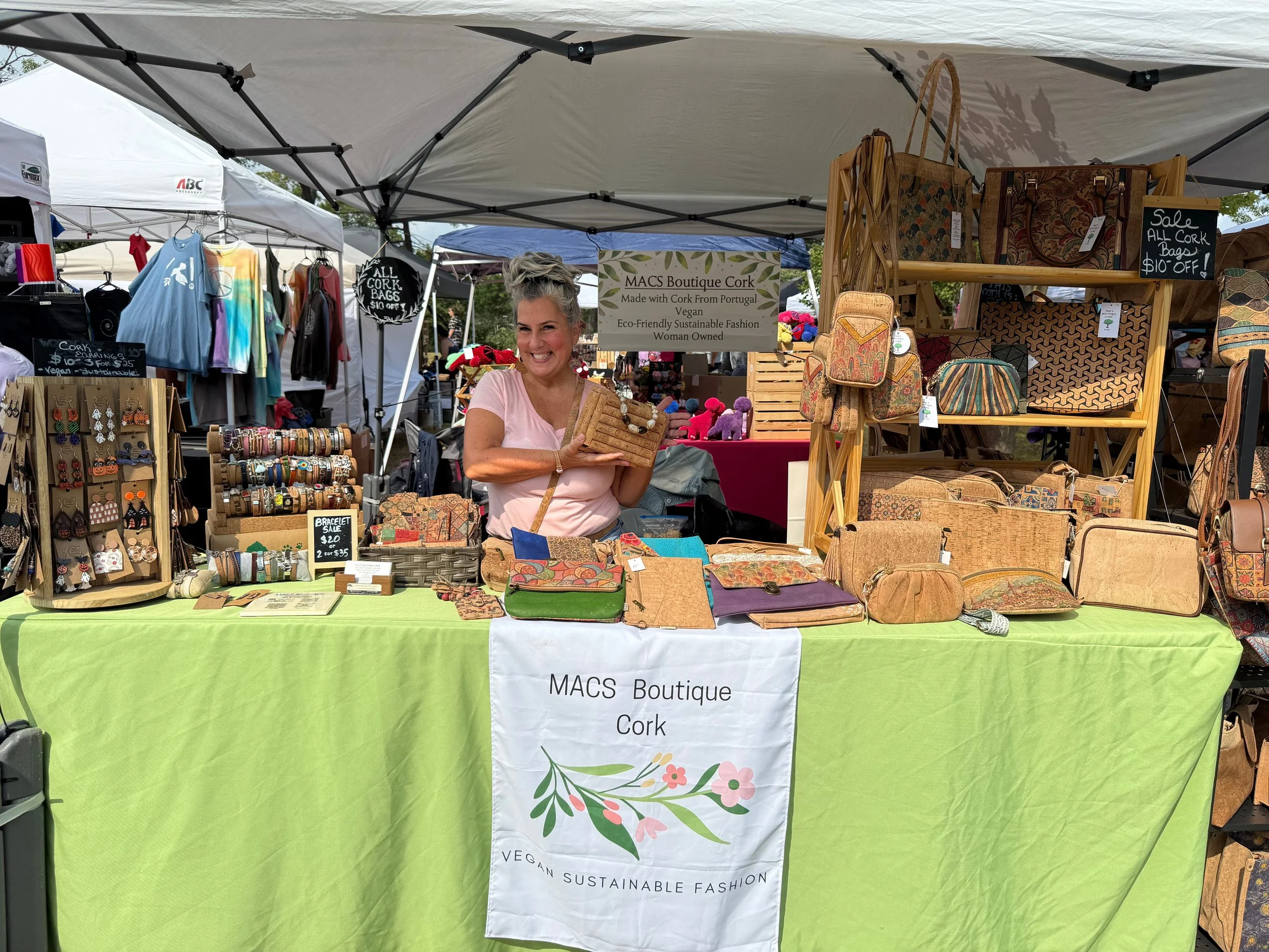 At an outdoor market stall, a woman with blonde hair tied up in a bun is smiling and holding a cork bag. The stall displays various cork products, including handbags, wallets, and earrings. There are signs indicating sales and product details. The backdrop shows other market stalls and trees.
