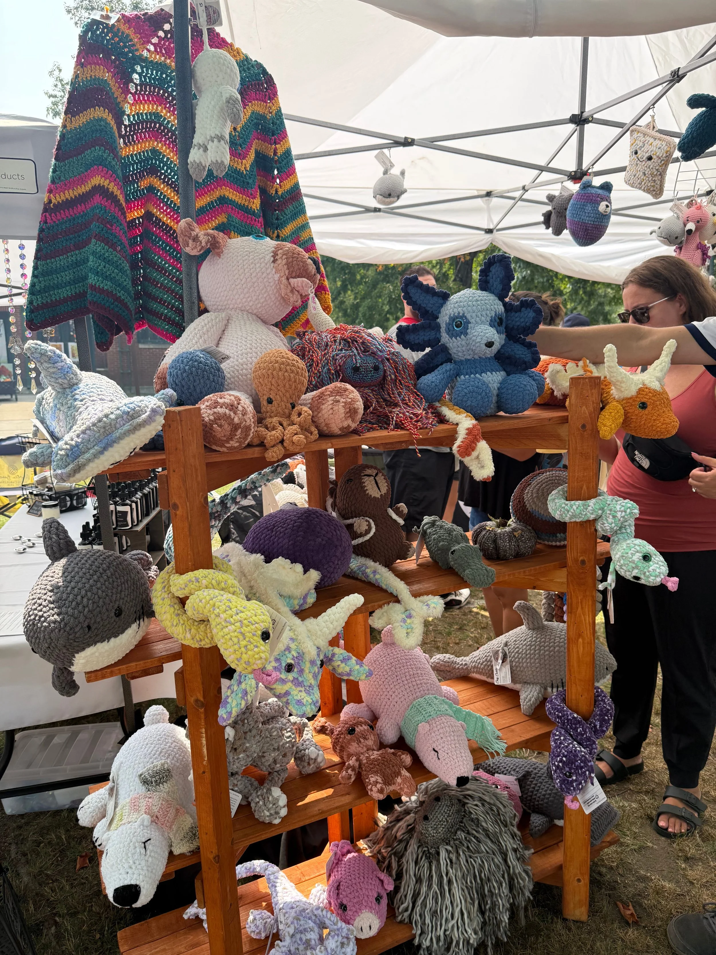 A display of handmade crocheted stuffed animals and hats at an outdoor market stall, including a wolf, unicorn, lion, and other animals, under a white tent.