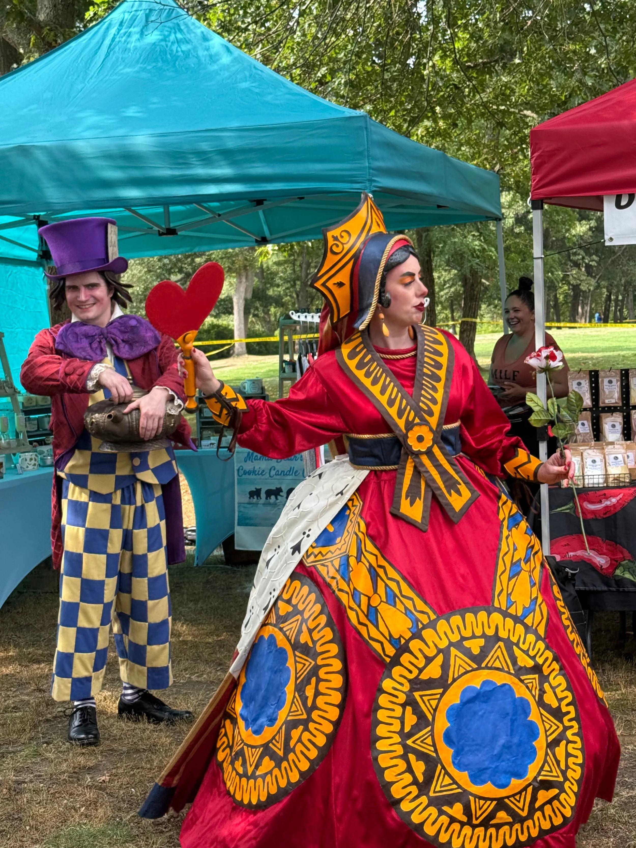 Two individuals dressed in colorful costumes standing under tents at an outdoor event. One person wears a predominantly red and yellow outfit with a large cloak and headpiece, while the other is dressed in a purple top hat and a purple scarf with yellow checkered pants. There are tables with various items behind them, and two women are smiling in the background.