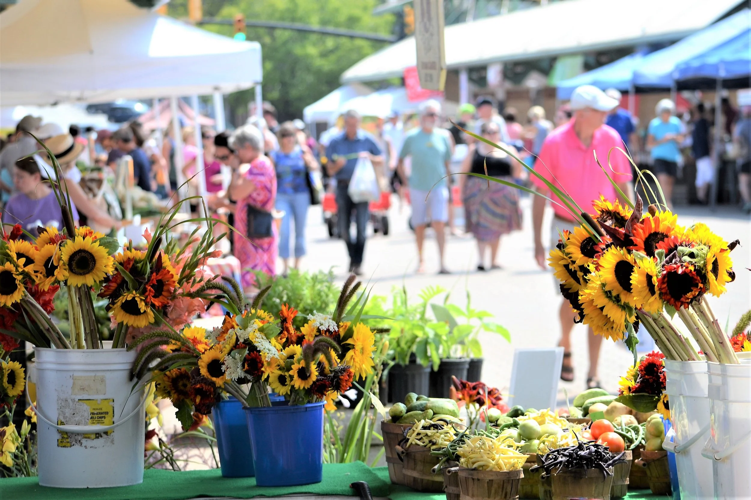 At an outdoor market, a table displays buckets of sunflowers and other flowers, with various vegetables including tomatoes, peppers, and eggplants visible on the table. People are walking and browsing in the background under tents and trees.