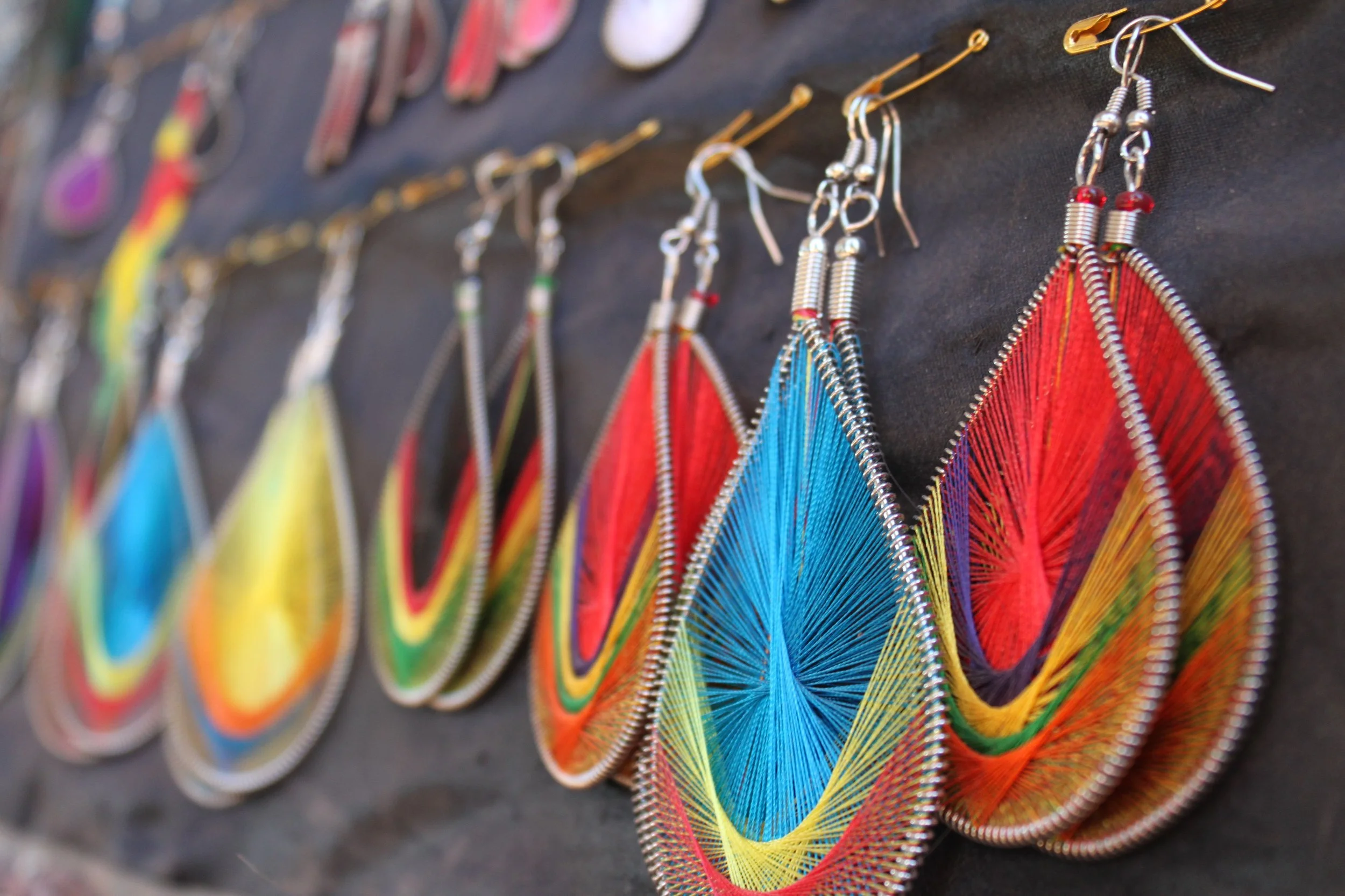 Colorful earrings hanging on a display. Each earring features thread work in rainbow colors, with silver coil accents and small beads, arranged in a fan-like pattern.