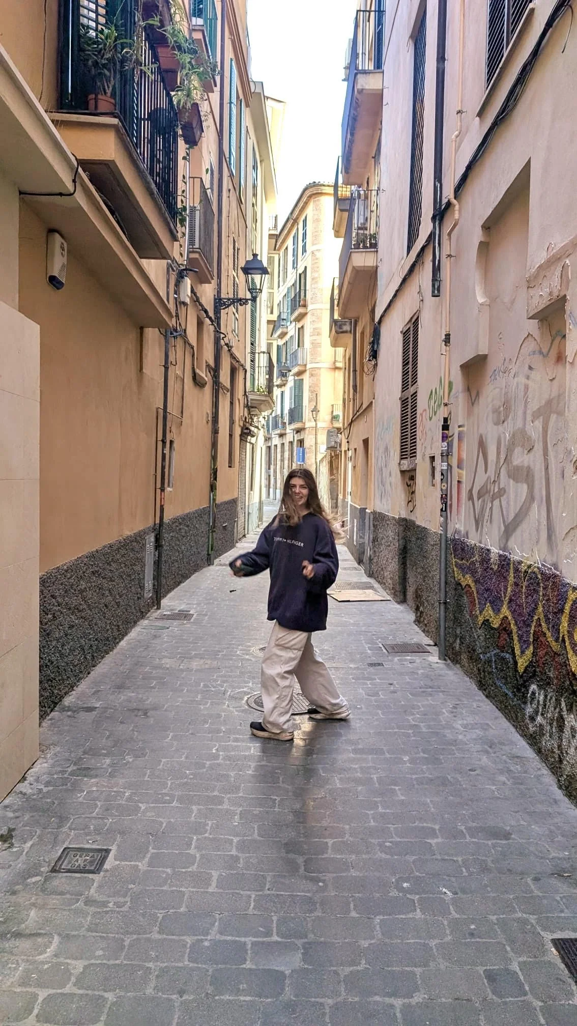 A person with long hair, smiling, standing in a narrow alleyway with buildings on either side, some with balconies and graffiti on the walls.