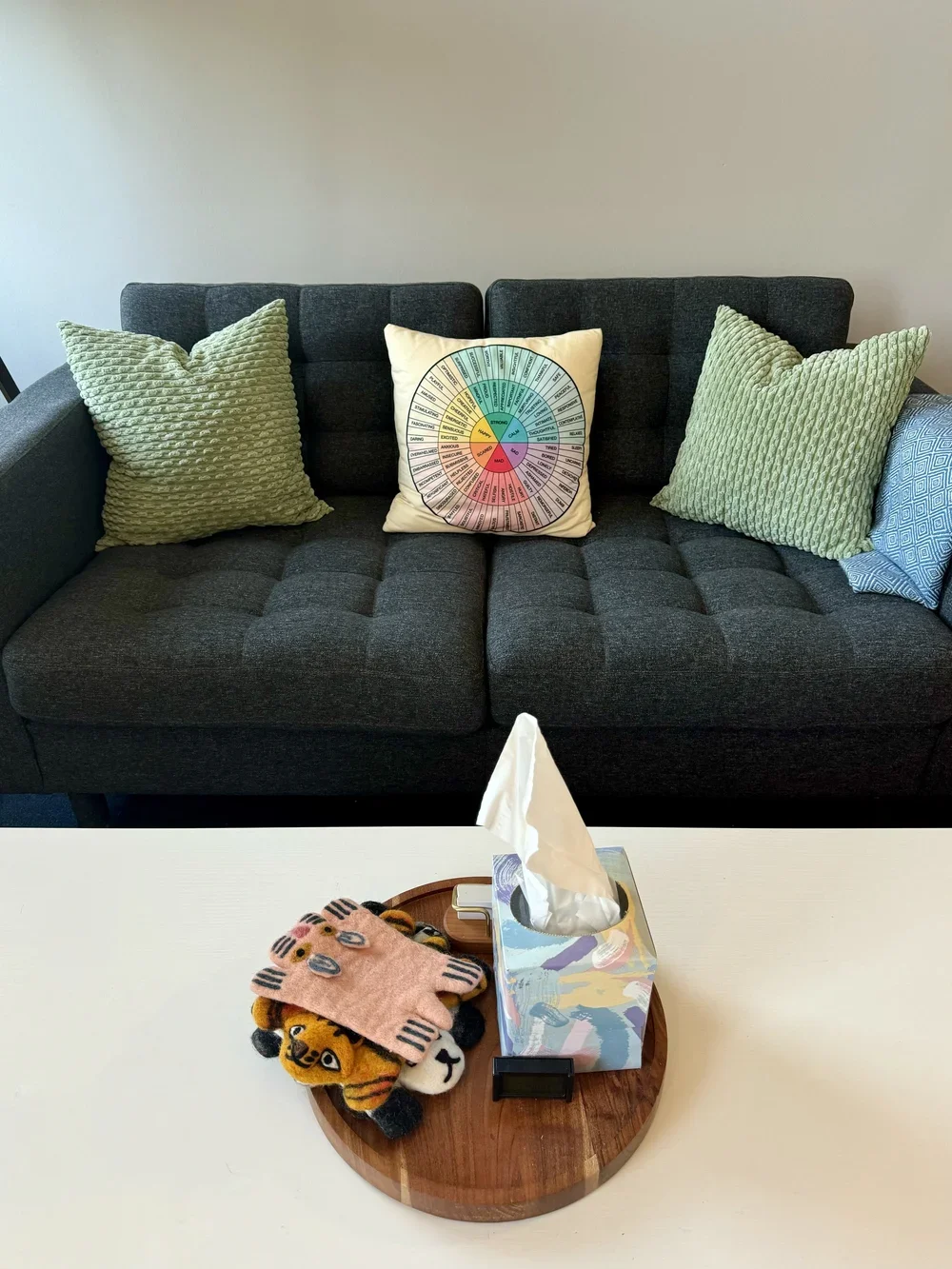 A dark gray sofa with three pillows, including two green textured pillows and a white pillow with a circular color chart, in front of a plain wall. In the foreground, a white table with a tissue box and small plush tiger toys on a round wooden tray.
