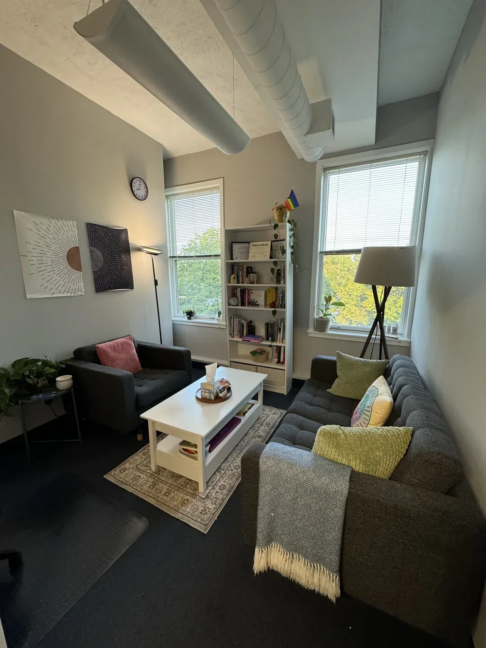 A cozy living room with a gray sofa adorned with yellow and patterned pillows, a black armchair with a pink pillow, a white coffee table, a bookshelf with decorations, artwork on the wall, and large windows letting in natural light.