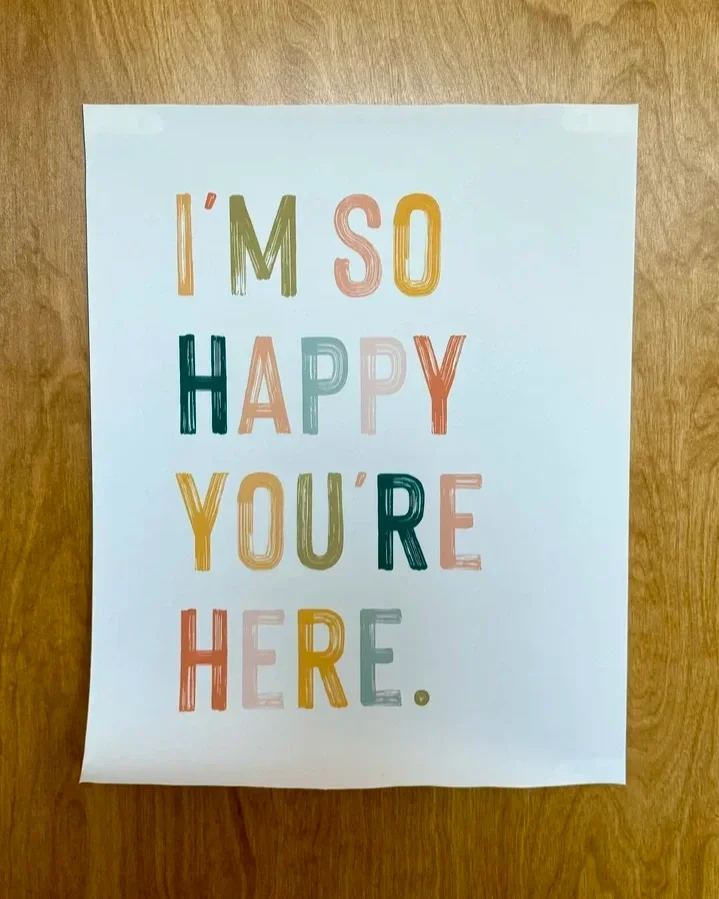Colorful printed sign on white paper that reads 'I'M SO HAPPY YOU'RE HERE.' in multicolored letters, taped to a wooden surface.